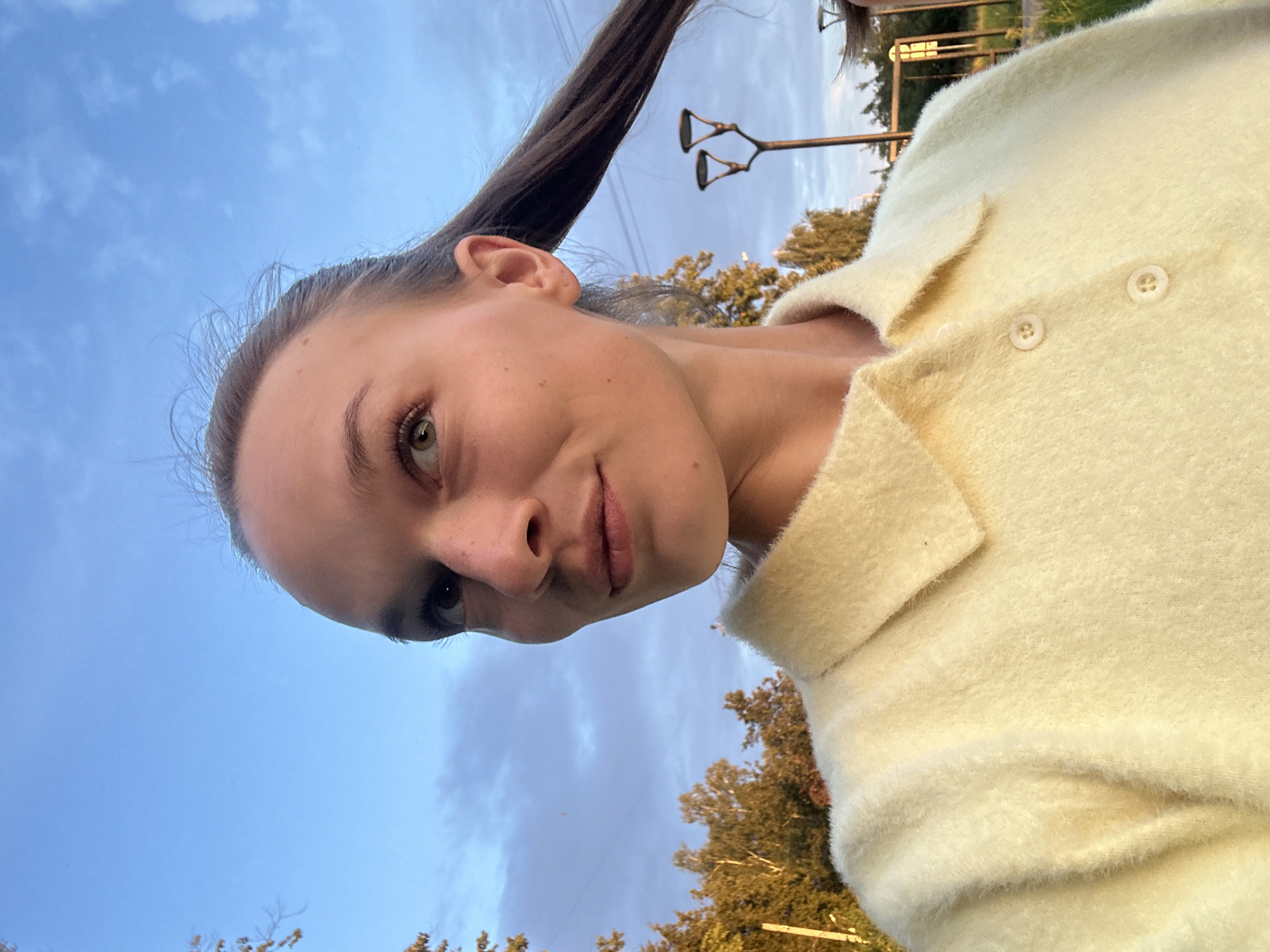 A young woman with light brown hair pulled back into a ponytail is taking a selfie outdoors. She is wearing a light yellow, fuzzy collared shirt. Her eyes are looking up and to her right, and she has a slight smile on her face. The background shows a blue sky with scattered clouds, trees with autumn foliage, and two streetlights. The scene is likely in Chaikovsky, Russia, given the context.