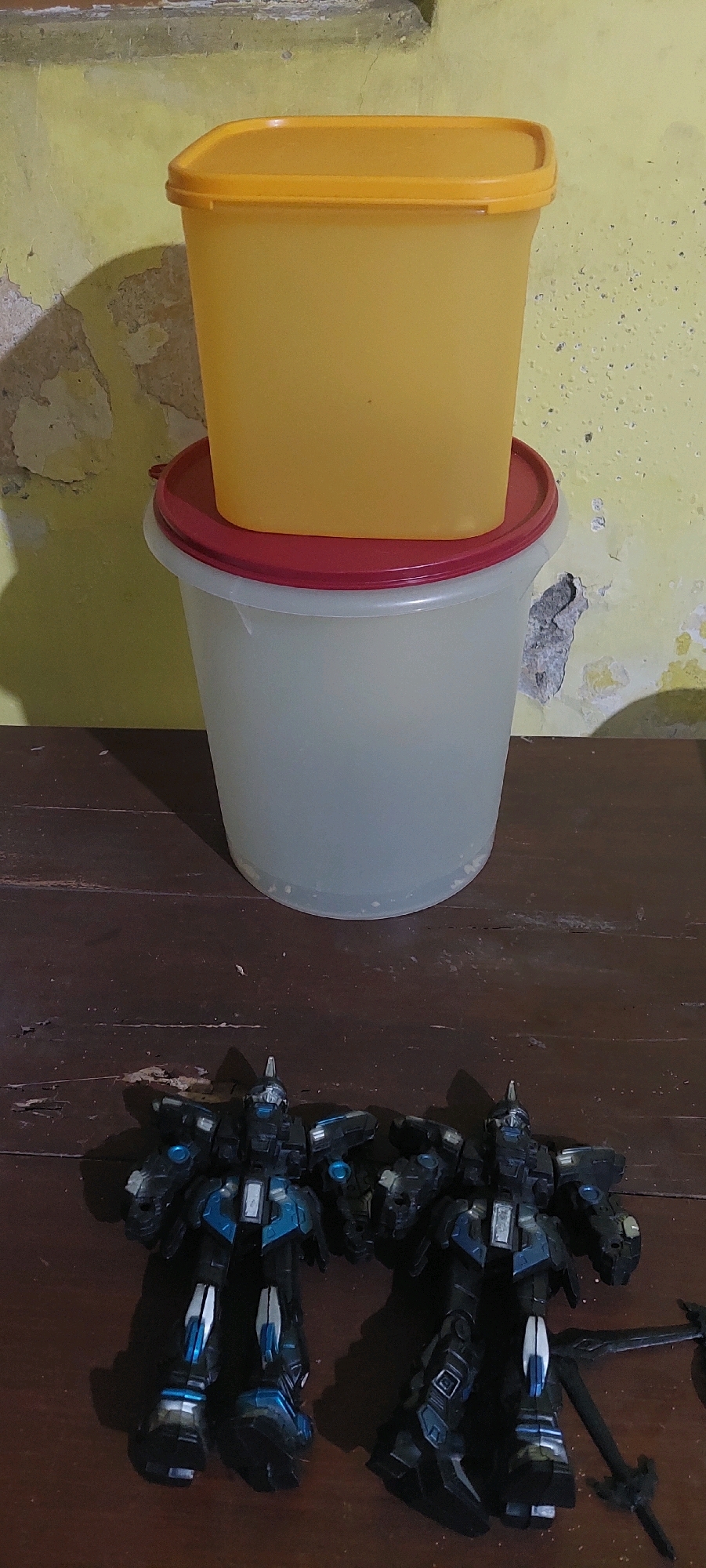 Two dark-colored, humanoid action figures with blue accents are positioned on a dark wooden surface. To the left, an action figure stands facing forward, its left arm extended. To the right, a second action figure is also facing forward, with its right arm extended. Behind the action figures, a large, translucent cylindrical container with a red rim is visible. Resting on top of the red rim is a yellow rectangular container with a matching yellow lid. The background consists of a textured, yellowish wall with patches of exposed concrete. The scene is lit from above, casting shadows from the objects onto the wall and surface.