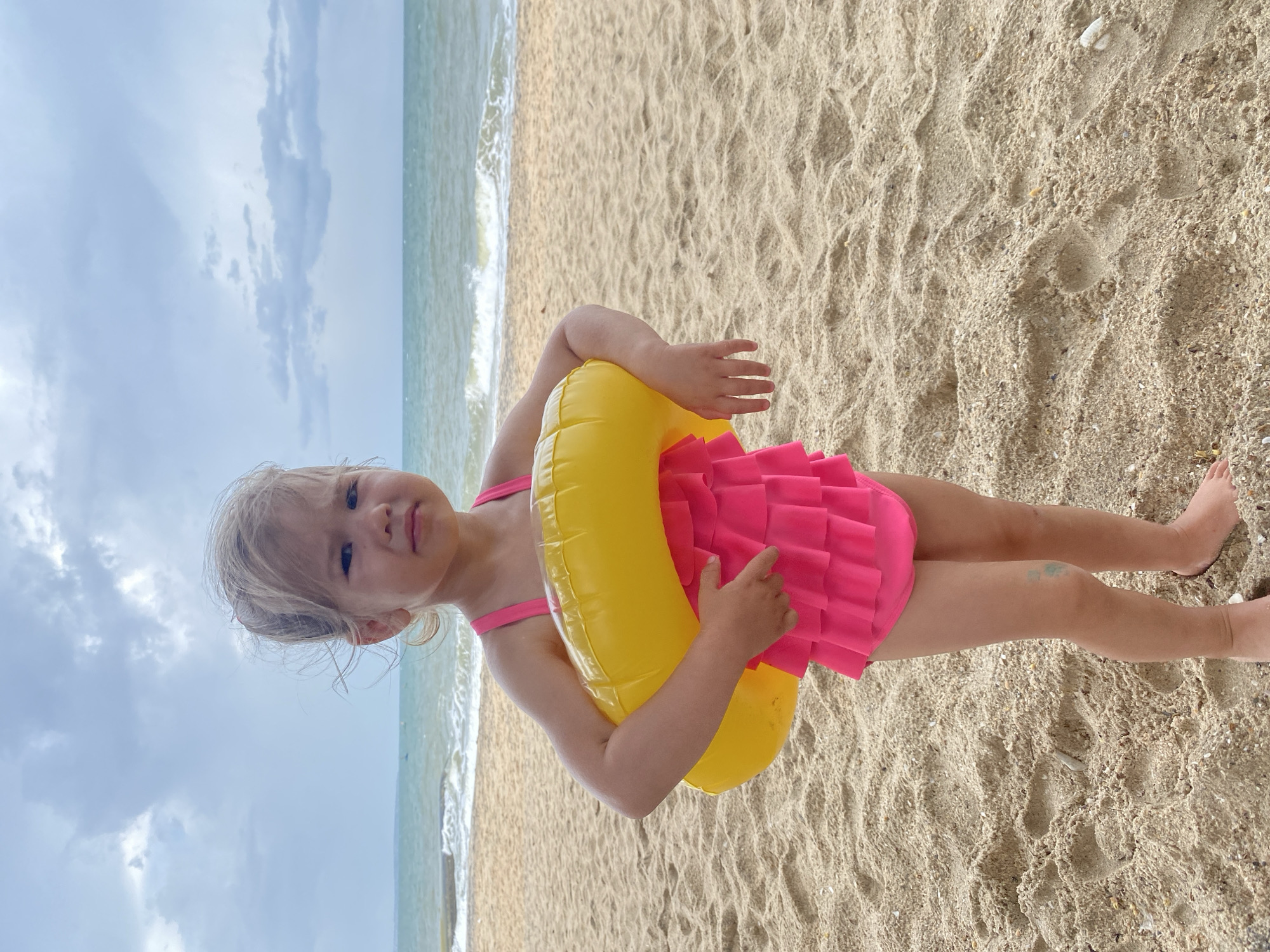 A young child, likely a girl with blonde hair, stands on a sandy beach, looking directly at the camera with a neutral expression. She is wearing a bright pink ruffled one-piece swimsuit and holds a yellow inflatable swim ring around her waist. Her bare feet are visible on the sand, which shows footprints and small shells. A faint greenish mark is visible on her left knee.
In the background, the ocean stretches out under a cloudy sky. Gentle waves break on the shore to the child's right. The scene depicts a relaxed moment at the beach, possibly just before or after swimming, on an overcast day. The setting is Phu Thuy Ward, Vietnam. There is no visible text in the image.