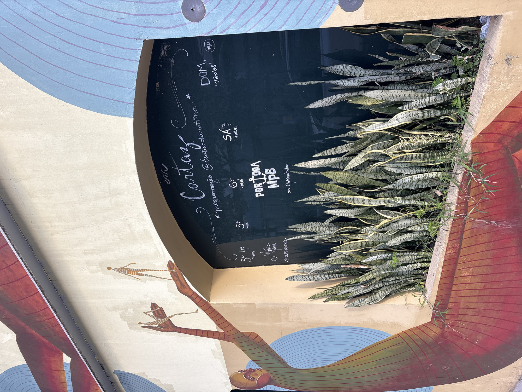 The image shows a mural on the exterior wall of a building. The mural features a stylized human figure on the left, with arms outstretched, and two smaller stick figures above. The right side of the mural includes an arched chalkboard with text written in white chalk. In front of the chalkboard, there are several snake plants growing in a planter. The text on the chalkboard includes "em Cartaz", "@teatrodarotina", and details about programming for different days, such as "DOM. 'Todos' 19H", "SÁB. 'todos'", and "6ª 'todas'". There is also text that appears to be "POR TODA MPB com Antônio Porto". The mural and the chalkboard suggest this is an event space or a cultural venue. The architecture of the building, with its arched opening and stucco walls, is typical of older constructions in São Paulo. The specific location is unknown from the image alone, but the style of the mural and the presence of Portuguese text suggest it is in Brazil.