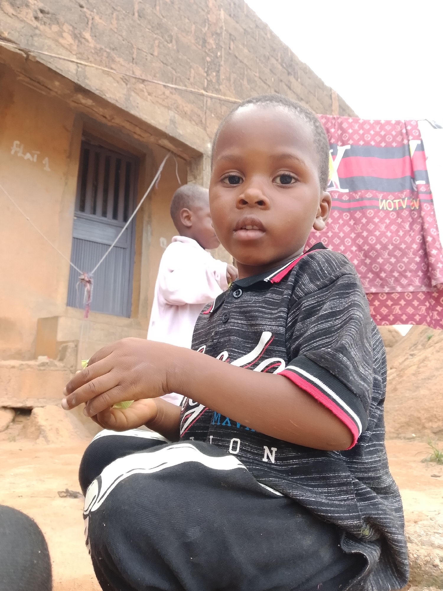 In an outdoor setting, a young Black child with short dark hair and brown eyes is seated in the foreground, looking directly at the camera. The child is wearing a black polo shirt with thin horizontal white stripes and a red and white striped collar, and black pants with a white stripe running down the side. The child's right hand is raised, holding a small green object, possibly a leaf. In the background, another child, also Black, is partially visible, wearing a light pink garment. Behind them, a textured, earthy-toned building with a grey barred window and a stone staircase is present. A patterned fabric, predominantly red and black with a repeating logo-like design, is hanging on a line to the right of the building. The ground is composed of reddish-brown dirt and rocks. The overall environment appears to be a rural or developing urban area.