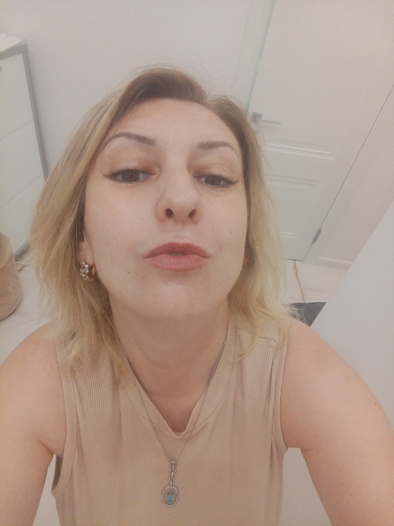 A woman with blonde hair is captured in a selfie. She is wearing a beige, sleeveless, ribbed tank top with a silver necklace that has a pendant. She has noticeable eyeliner and what appears to be mascara on her eyelashes. There is a stud earring on her right ear. Her lips are slightly pursed. The background suggests an indoor setting, possibly a bathroom or a room with light-colored walls and doors, with what might be a reflective surface on the left, possibly a mirror or a closet door. No specific location in Sochi, Russia, is indicated.