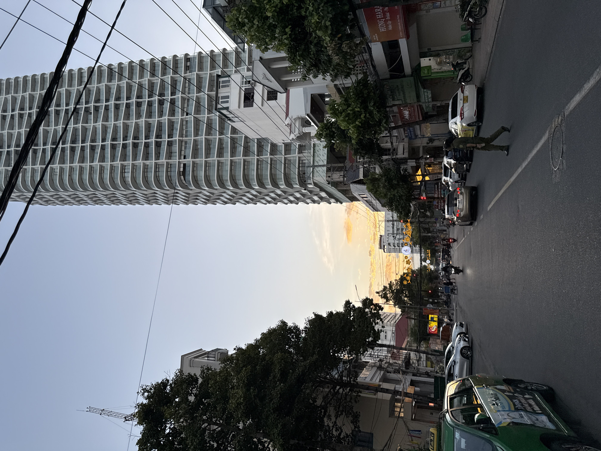 The image captures a street scene in Nha Trang, Vietnam, during what appears to be dusk or early evening. A prominent, modern, multi-story building with a distinctive wavy facade dominates the right side of the frame, its numerous balconies giving it a unique texture. Utility wires crisscross the sky in the foreground, partially obscuring the building and the sky.
The street itself is paved and shows signs of moderate traffic. Several vehicles, including cars and motorcycles, are visible. A green taxi is parked on the left side of the street, displaying advertisements on its side. A person in what appears to be a uniform is walking across the street from right to left in the middle ground. On the right side of the street, buildings line the road, some with shops or businesses displaying signage, including one with the text "HUNG HARA Make Up - Bridal - Ph". Trees with dark green foliage are interspersed along the sidewalks and near the buildings.
The sky exhibits the warm hues of a sunset, with patches of orange and yellow clouds against a light blue background. Streetlights and some building lights are beginning to illuminate, indicating the transition from day to night. The overall atmosphere suggests an urban environment bustling with activity as the day winds down.