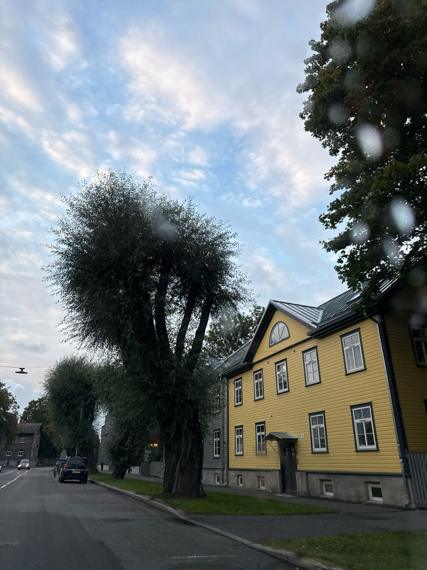 The image captures a street scene in Tallinn, Estonia, likely during late afternoon or early evening, given the soft lighting and cloud cover. The focal point is a bright yellow wooden house with a metal roof, featuring multiple white-framed windows and a small covered entranceway. Adjacent to this house are large, mature trees with dense foliage, one of which is particularly prominent and appears to be a willow.
On the street, a black SUV is parked, and a few other cars are visible further down the road, suggesting moderate traffic. The street itself is paved, with a sidewalk and a strip of grass separating the road from the houses and trees. The overall environment appears to be a residential or mixed-use area within the city. The sky is a mix of blue and white clouds. Some blurred circles on the foreground suggest the image was taken through a wet window, possibly due to rain.