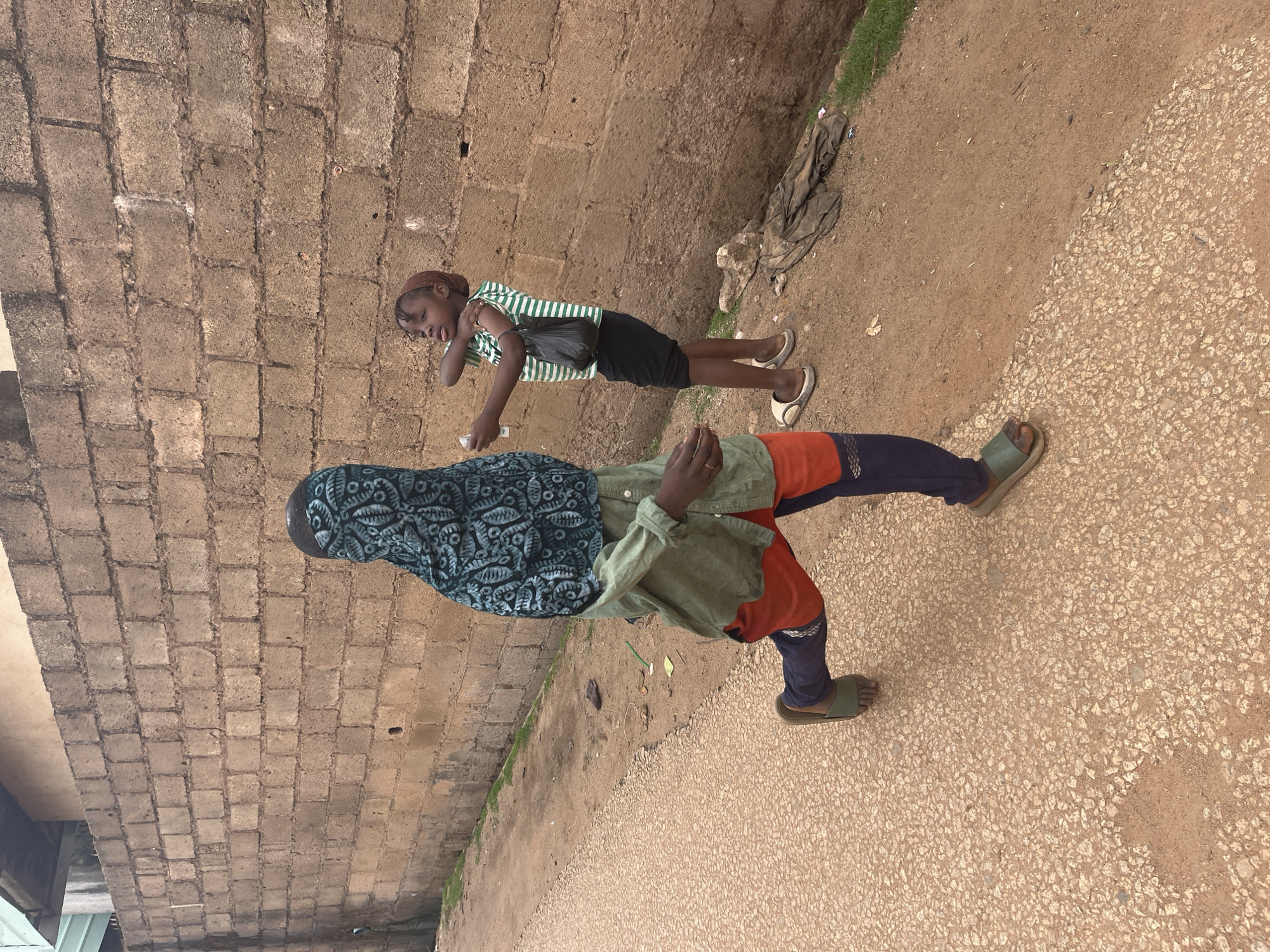 The image depicts two young individuals on a dusty street in Jos, Nigeria. On the left, a person wearing a patterned headscarf that covers their face and neck, a loose-fitting olive green long-sleeved shirt, and bright orange and navy blue trousers is in motion, with their left leg extended forward and right leg back, suggesting they are walking or running. They are wearing green slides on their feet. In the center-right of the image, a second individual, appearing to be a girl, stands facing the left. She has short hair and is wearing a brown head covering. Her outfit consists of a green and white striped sleeveless t-shirt and a black skirt. She holds a black plastic bag in her left hand and appears to be gesturing with her right arm, possibly reaching towards the other person, who is holding a white object, likely a bottle, in their right hand. The background is dominated by a rough, brown brick wall. The ground is a mix of a rough, speckled asphalt-like surface on the left and dirt on the right, with some scattered debris and grass near the wall. The lighting suggests an overcast day.