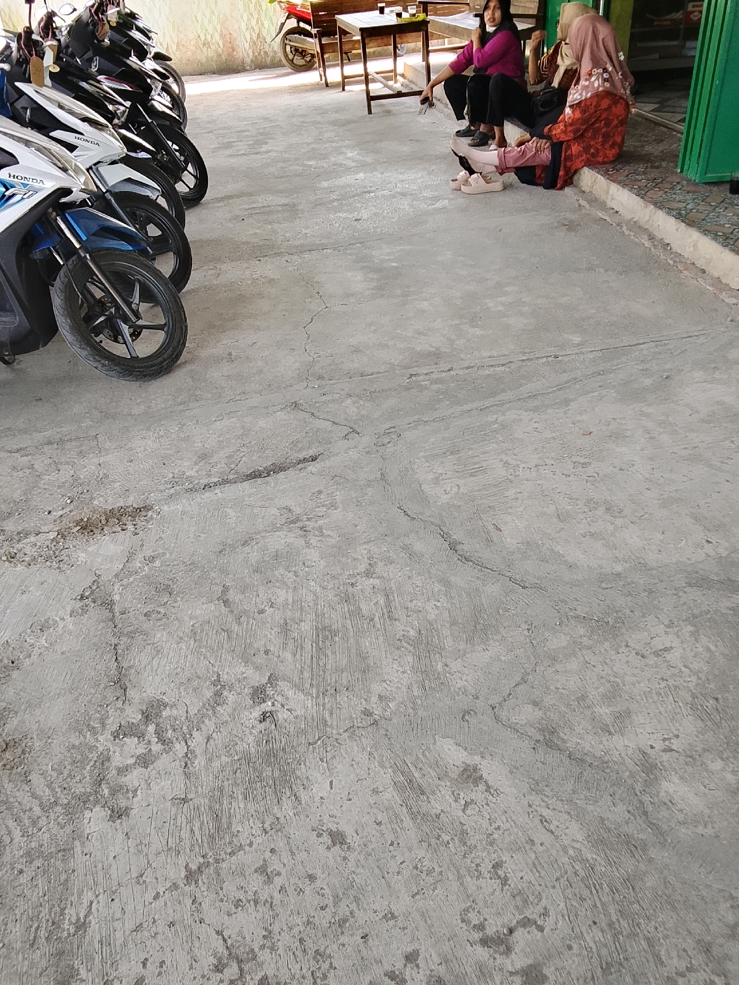 In Tayem, Indonesia, a group of individuals is seated on a concrete curb, facing away from a line of parked motorcycles. The motorcycles are all facing the same direction, parked neatly in a row on a paved surface. The environment appears to be an outdoor area, possibly a roadside or a parking lot, with a building or wall in the background. The people are casually dressed, with two women wearing headscarves and one woman in a pink top and black pants. The overall scene is one of relaxation or waiting.