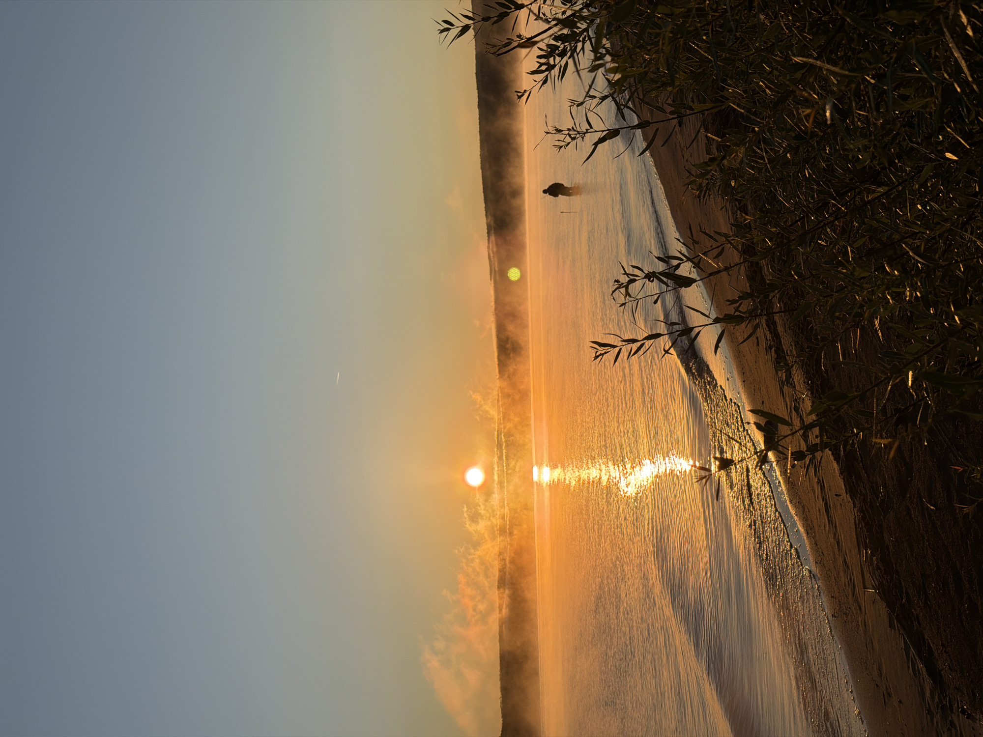 This is a vertical, outdoor, daytime, vertical photograph depicting a serene sunrise over a calm body of water, likely a reservoir or lake, adjacent to a sandy shore. The sun, a bright orb of orange, is positioned in the upper left quadrant, casting a golden, reflective sheen across the water's surface. Wisps of mist or fog hang low over the water, illuminated by the sun's rays, creating a soft, diffused glow. In the mid-ground, a lone figure stands in the shallow water, silhouetted against the bright sky, appearing to be fishing or wading. The shoreline in the foreground is composed of sand and features dark, out-of-focus foliage in the lower right corner, suggesting it is a natural bank. The sky transitions from a pale blue at the top to a warm orange and yellow gradient near the horizon. The overall atmosphere is tranquil and evokes a sense of early morning peace.
The location is identified as the Kama River reservoir near Chaikovsky, Russia.