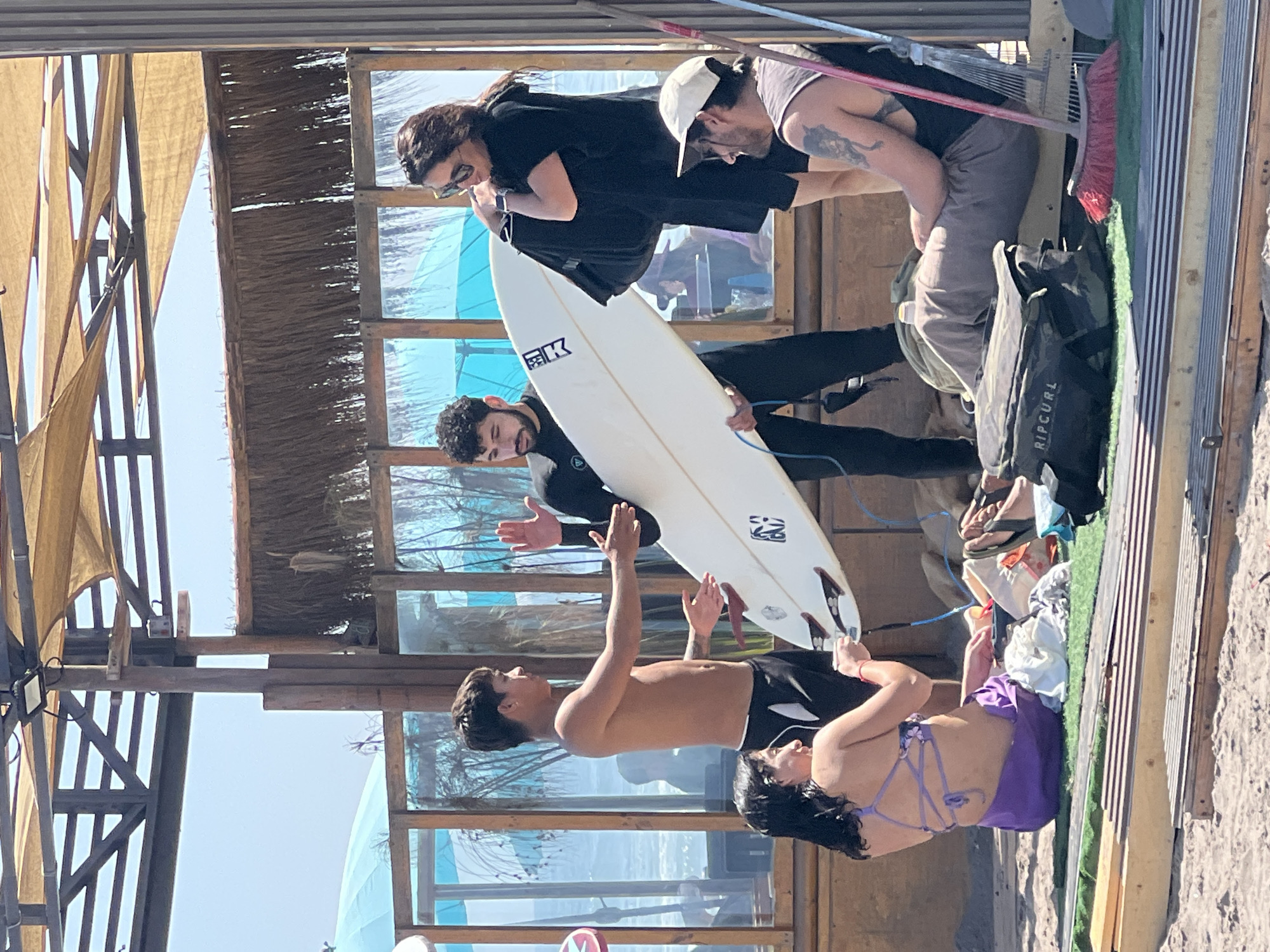 The image captures a scene on a sunny day in what appears to be a coastal area, possibly in Arica, Chile. Four individuals are visible: a shirtless man with dark hair standing, his arms raised, possibly interacting with a white surfboard held by another person in a black wetsuit; next to the shirtless man, a woman in a purple bathing suit and dark hair leans back; two individuals are above and behind them, a woman in sunglasses and a black shirt looking down at the surfer, and another person with a white cap and visible tattoo is behind her, looking down at the scene. The people are situated in a wooden, semi-open structure near a beach, with blue-tinted windows in the background showing a glimpse of the ocean. A "Ripcurl" brand bag and some clothes are on a wooden platform next to the seated woman. The structure appears to be part of a building at or near the beach.