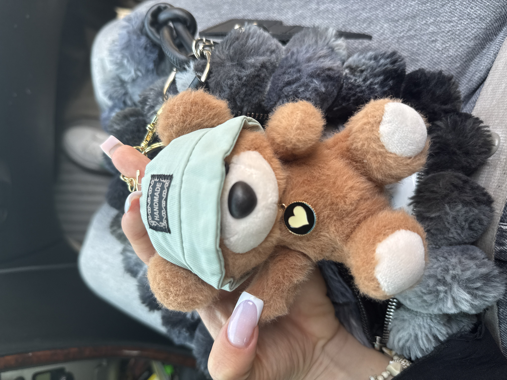 The image displays a close-up shot of a person's hand holding a brown plush teddy bear keychain inside a car, with the scene set in Sofia, Bulgaria. The teddy bear is soft and furry, featuring white-colored pads on its paws. It is notably wearing a light green face mask, which has a small black tag with the word "HANDMADE" printed in white. A tiny, circular black charm with a yellow heart is attached near the teddy bear's ear.
The hand holding the keychain has manicured nails, adorned with a classic French tip style. In the background, a fuzzy accessory or bag covered in dark grey and black pom-poms is visible, partially obscured. Parts of the car's interior, including what appears to be a dashboard or console on the left and grey fabric upholstery, can be seen, confirming the setting. The lighting suggests it is daytime. The primary activity is the holding of the keychain, with no specific event or strong emotions discernible.