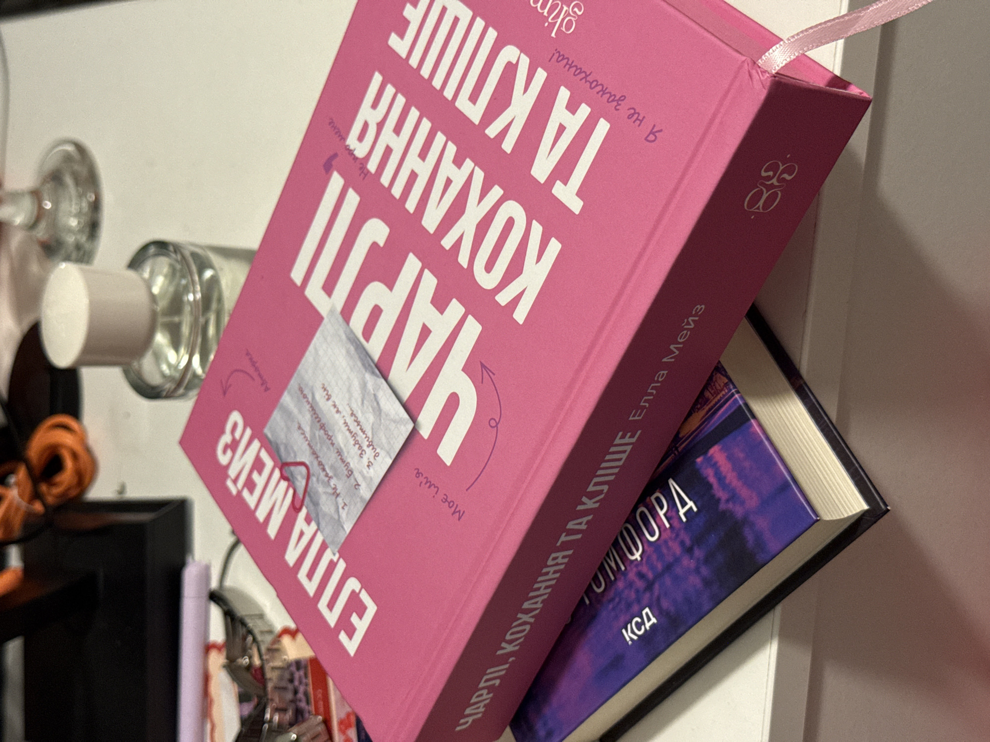 The image shows a close-up of two books on a white surface. The top book is bright pink and has large white lettering that reads "ЧАРЛІ, КОХАННЯ ТА КЛІШЕ" and "ELLA MEIZ". A small, crumpled piece of paper with handwritten text is attached to the pink book with a red paperclip. The text on the paper appears to be a numbered list. The second book, positioned underneath the pink book, has a dark purple cover with text that is partially obscured. The background is blurred but suggests an indoor setting with some objects, including a glass bottle with a white cap and what appears to be a wine glass. There are no people visible in the image, and no specific location in Łódź, Poland, can be identified.