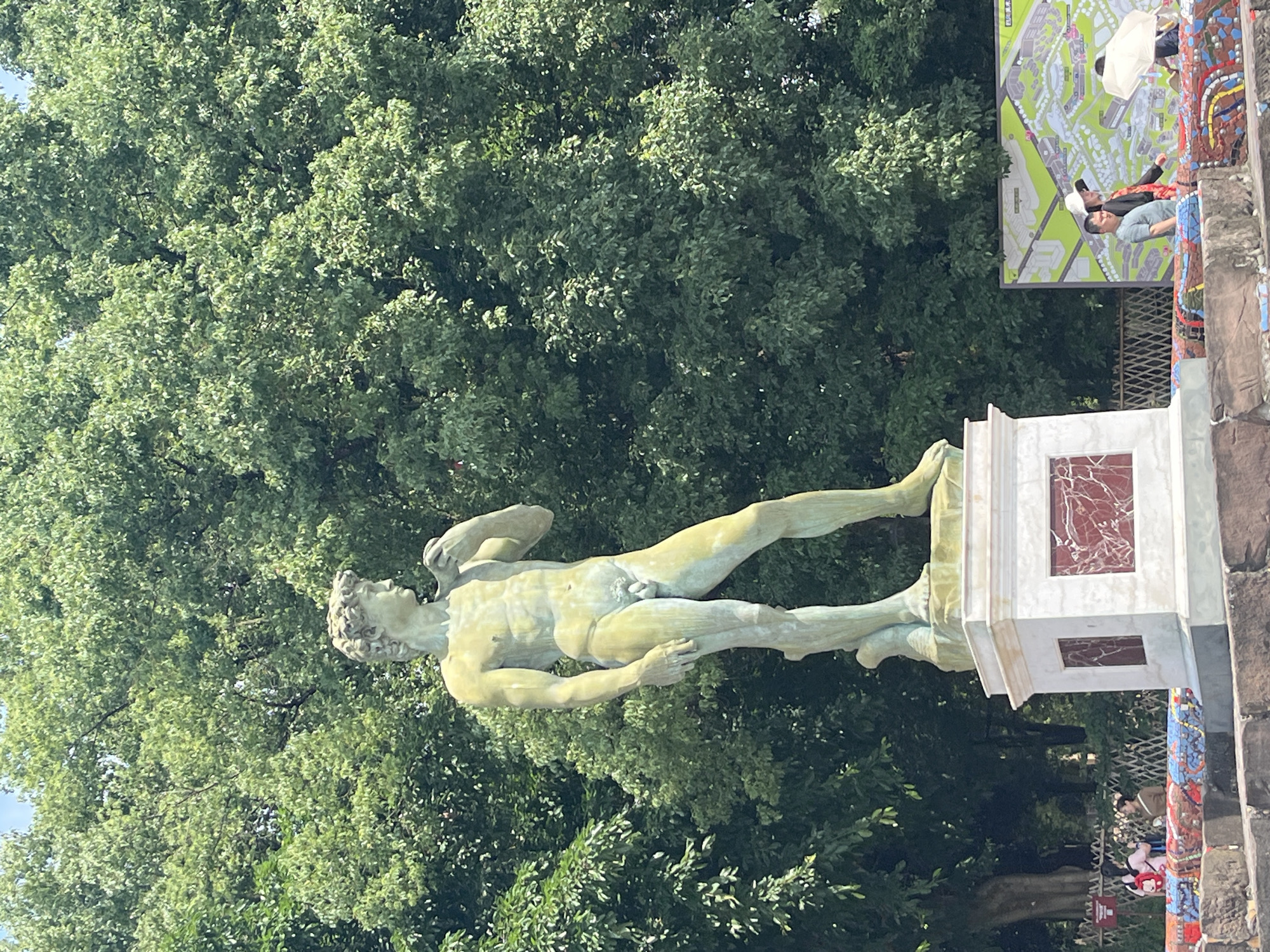 This image depicts a full-body statue of a male figure, resembling Michelangelo's David, standing on a white pedestal with red marble accents. The statue is outdoors, with a dense backdrop of lush green trees. In the lower right of the frame, a large map is displayed, with several people walking by it. One person is holding a white umbrella. The foreground shows a colorful, mosaic-like border. The scene appears to be in a public park or outdoor exhibition space. This image was taken in the High-tech Zone of Chongqing, China.