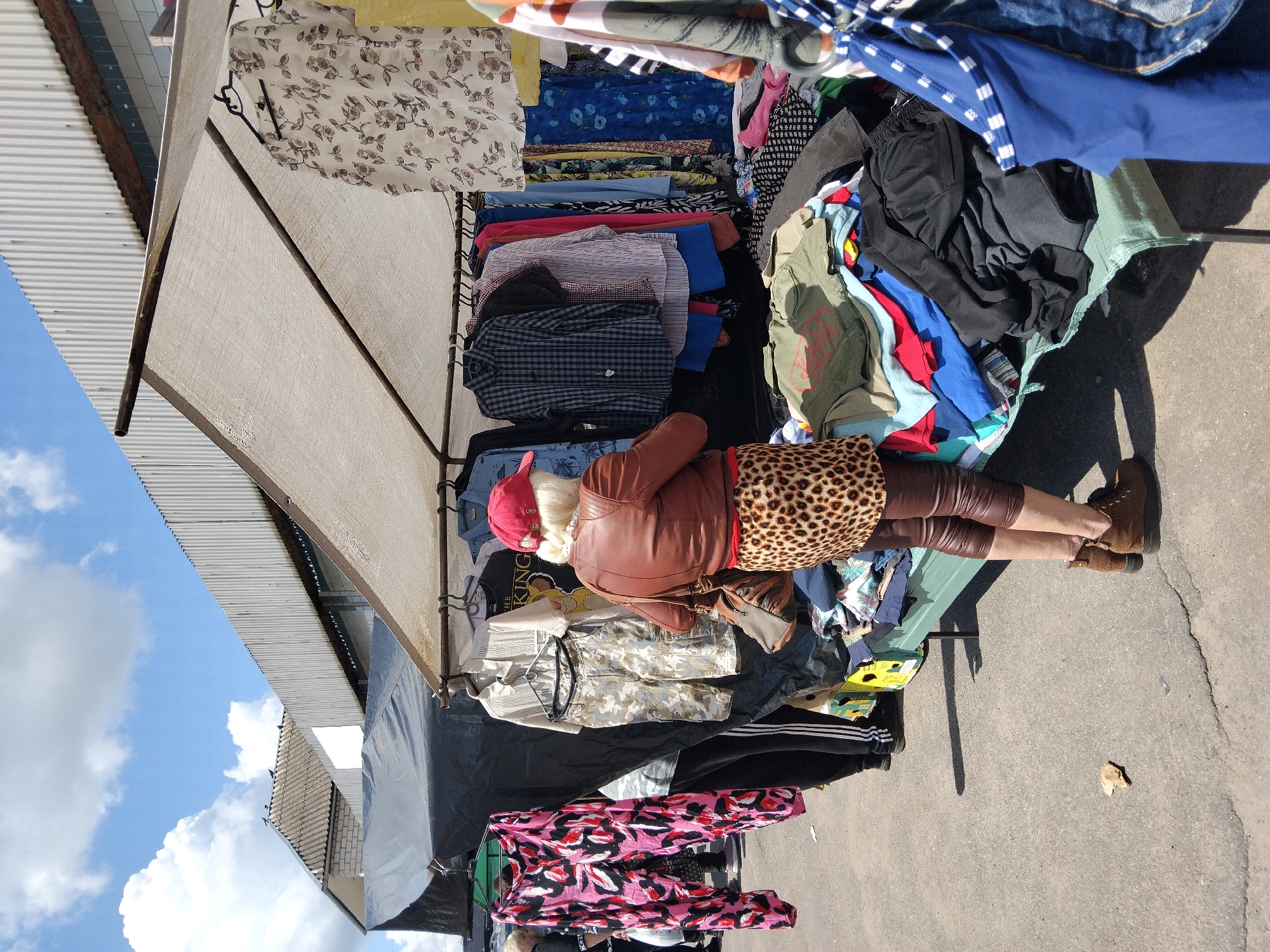 In Chudniv, Ukraine, an outdoor market stall is set up under a canopy. A woman, wearing a brown leather jacket, a red baseball cap, leopard-print skirt, and brown leggings, stands examining clothing. The stall displays a variety of garments, including shirts, pants, and a patterned dress, on hangers and piled on a table. The ground is paved, and the sky above is blue with white clouds.