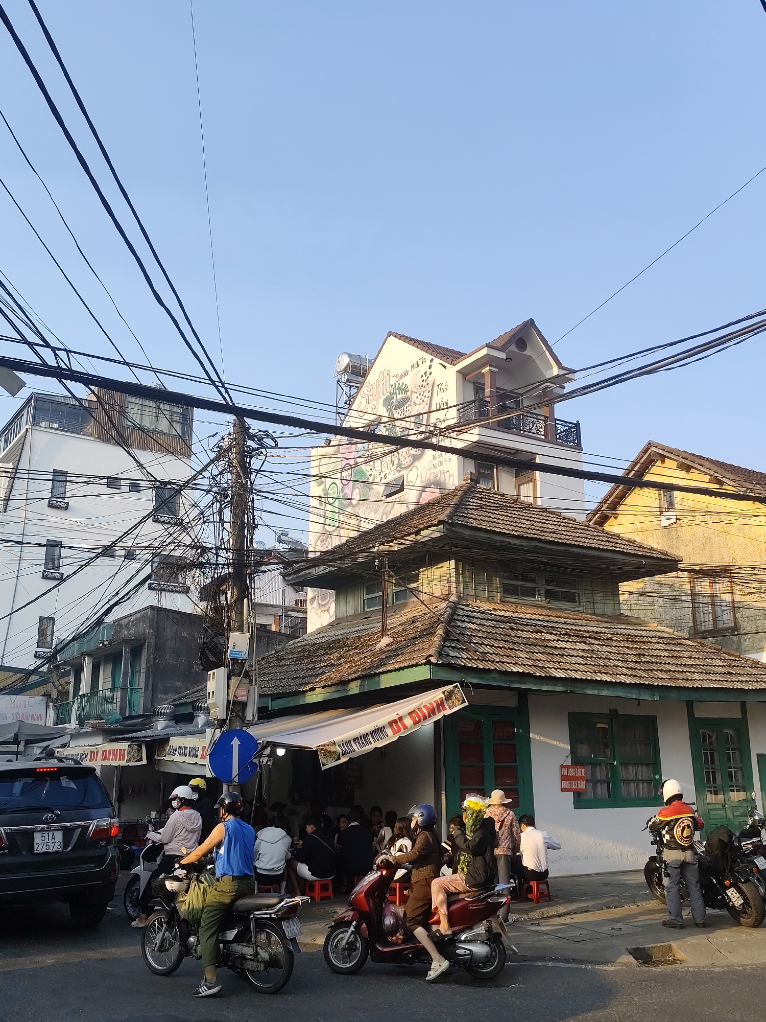 The image captures a street scene in Cam Ly - Đà Lạt, Vietnam. A complex network of overhead power lines dominates the upper part of the frame. Several buildings of varying heights and architectural styles are visible. One building is painted white with a mural featuring images of bubble milk tea, adding a touch of modern street art to the traditional setting.
In the foreground, a street-level view shows a lively scene. A mix of motorcycles and a black SUV are present on the street, and pedestrians are scattered around. Some people are seated on stools outside a building, likely a local eatery, under an awning that reads "DÌ DINH." The street-level activity indicates a bustling urban environment. There's a blue direction sign on the sidewalk to the left. The photo appears to be taken during daylight hours, based on the lighting.