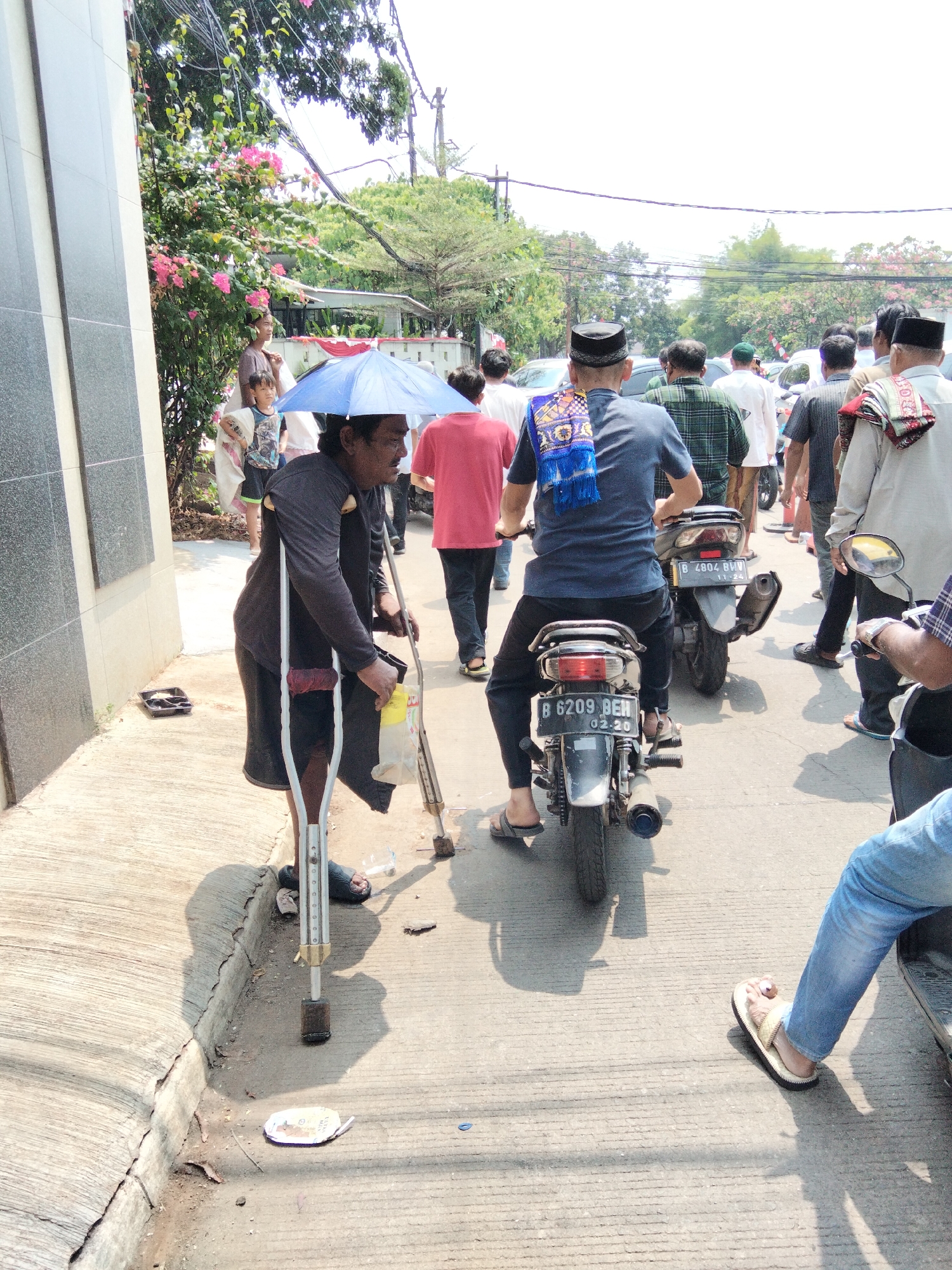 The image depicts a street scene in Depok, Indonesia, with a group of people and motorcycles. In the foreground, a man with crutches and an umbrella stands on the sidewalk. He is wearing dark clothing. Behind him, a person on a motorcycle, wearing a blue shirt and a patterned sash, is partially visible. Several other people, some wearing traditional attire, are walking or riding motorcycles in the background. The environment appears to be a public street with buildings and trees visible. The overall atmosphere is that of a busy urban area.