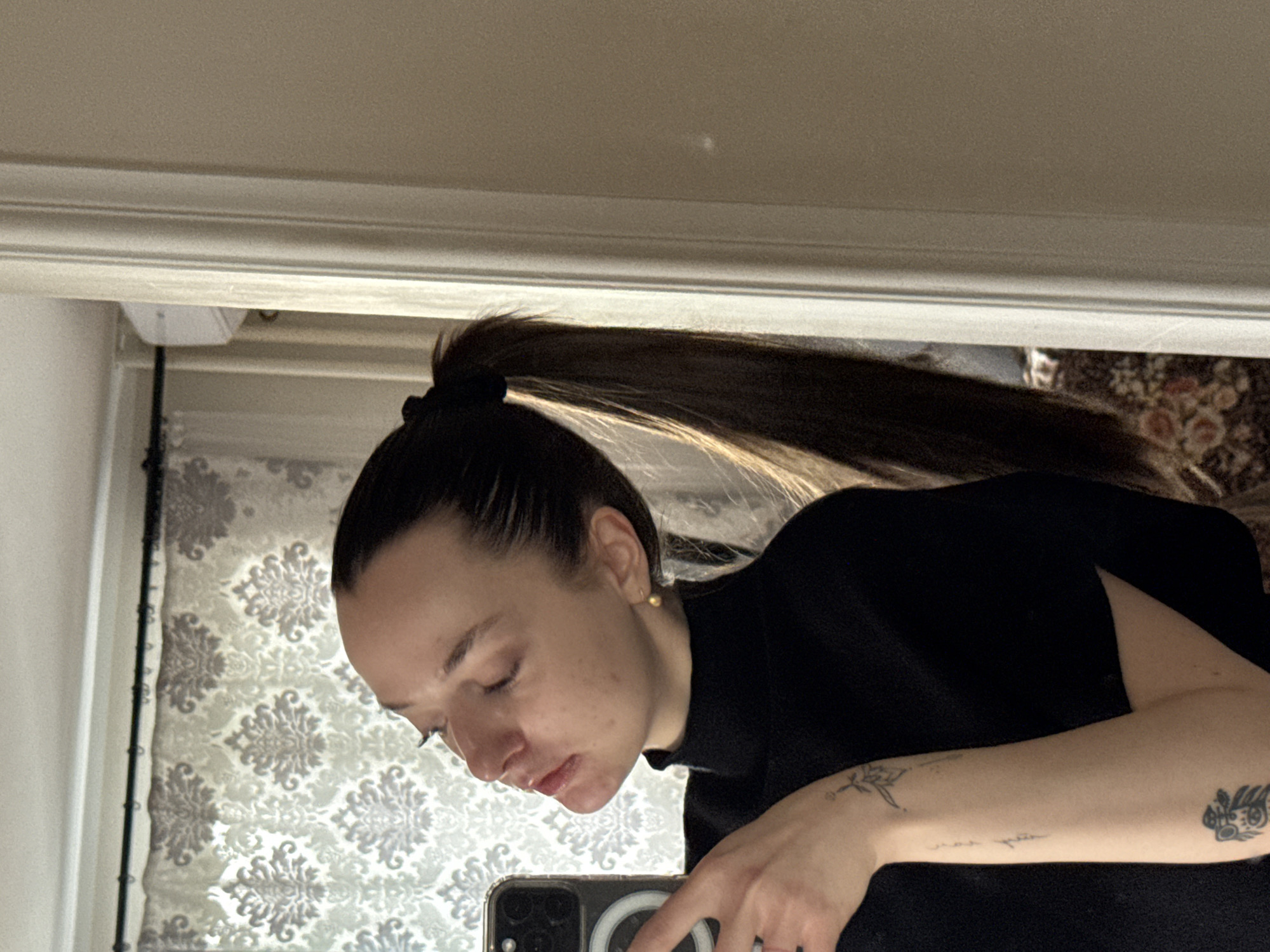 A woman with brown hair in a high ponytail is taking a selfie in a mirror. She is wearing a black turtleneck sweater. Tattoos are visible on her left arm. The background shows a wall with floral patterned wallpaper and a white door frame. The location is indoors and no specific location or reference to the city of Palilula Urban Municipality, Serbia is possible based on image analysis.
