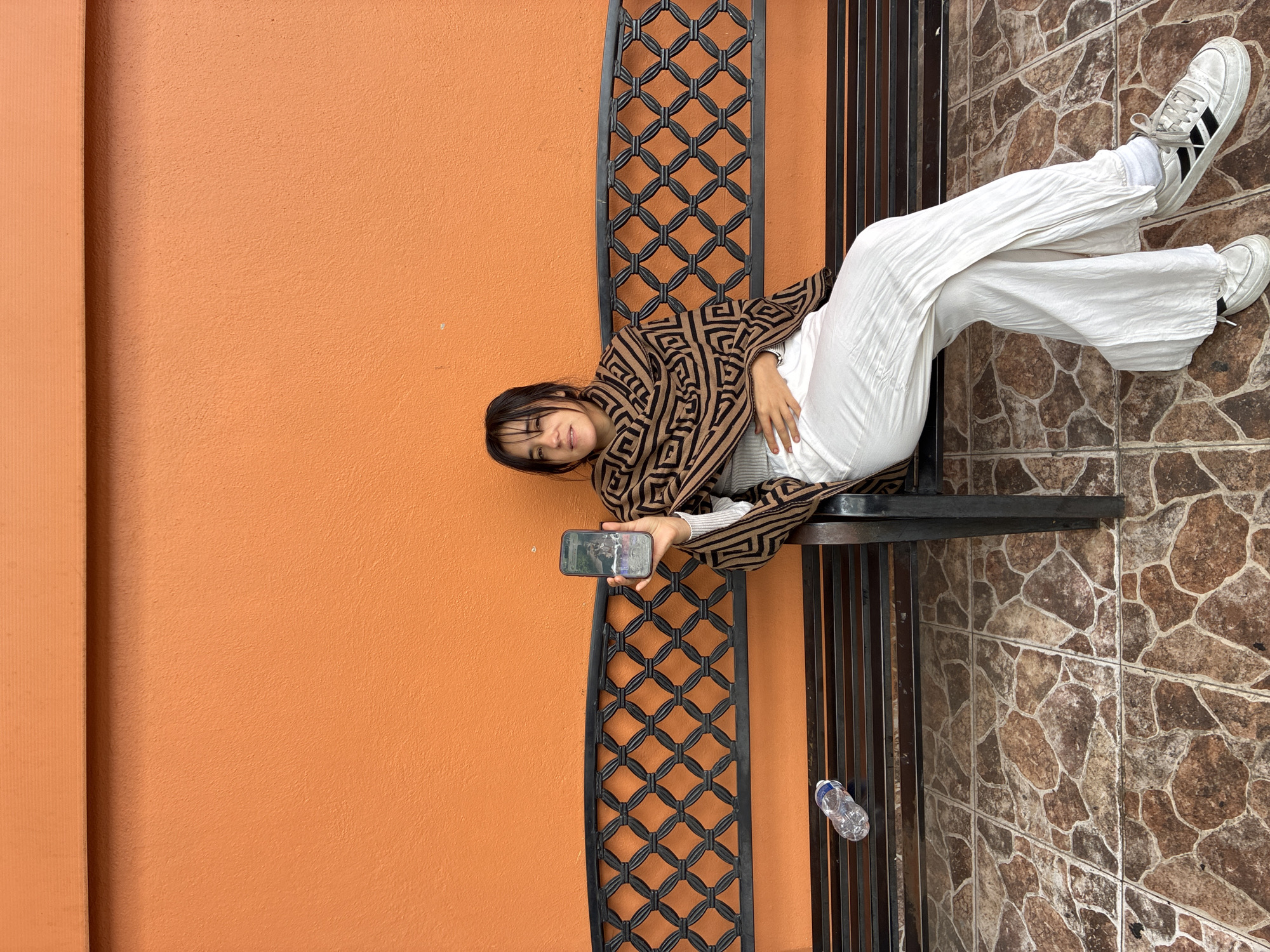 A young woman is lying on a bench in Huixquilucan de Degollado, Mexico, holding a smartphone in her hand, seemingly taking a selfie or watching something on the screen. She is wearing a brown and black patterned jacket, a white shirt underneath, and white pants. She has dark hair and is facing the camera. The bench is black metal with a decorative lattice pattern on the sides. Next to the bench, on the wall, there is a bottle on the ground. The background consists of an orange wall and a tiled surface.