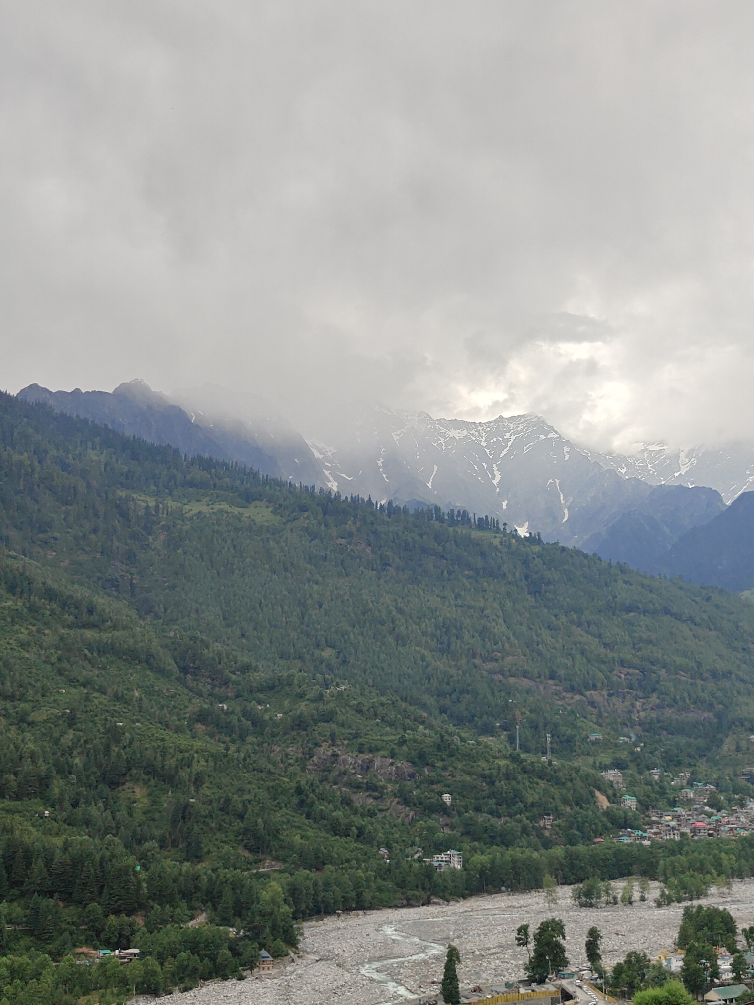 The image captures a wide, expansive view of a mountainous valley under a heavy, overcast sky. The upper portion of the frame is dominated by thick, light grey clouds, which partially obscure the distant peaks of a mountain range. Visible through the cloud breaks are snow-capped mountains, displaying white snow patches on dark grey rock faces.
Below the distant peaks, a prominent, steep hillside slopes downwards, densely covered in dark green coniferous and deciduous trees. Within this extensive forest, several scattered buildings, some appearing multi-story, are discernible, indicating human presence integrated into the natural environment.
In the mid-ground and foreground, a wide, rocky riverbed with a winding river occupies a significant portion of the lower frame. The riverbed is primarily composed of light grey and white stones and boulders. A stream of lighter-colored water flows through the center of the bed, consistent with the Beas River in the Kullu Valley.
On the lower slopes of the mountain on the right side of the river, a compact settlement or town is visible. This area features numerous buildings with varied roof colors, some clustered, and some more dispersed. Infrastructure includes visible roads with parked vehicles near the river in the bottom right, and at least two distinct communication towers (likely cellular or radio) are positioned on the forested slopes above the settlement. The entire scene is characterized by dense vegetation, rocky terrain, and human development interspersed within the natural landscape, indicative of a location such as Vashisht, India. No distinct human figures are visible in the image.