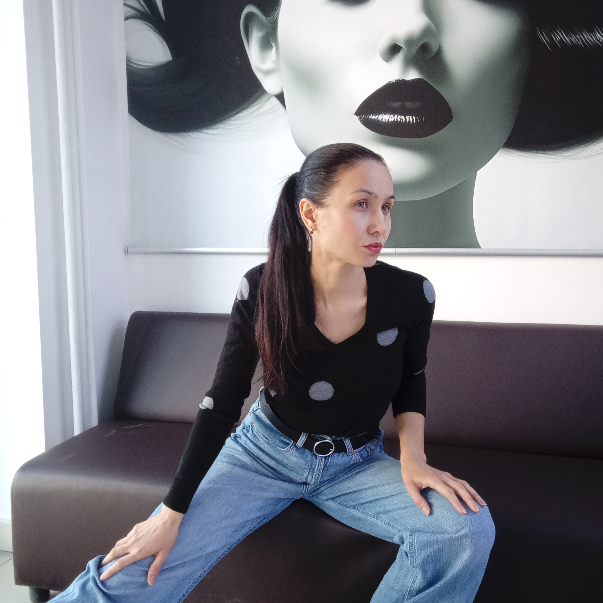 A woman sits on a dark brown couch with her legs spread. She wears a black long-sleeved shirt with grey polka dots and light blue jeans with a black belt. Her hair is pulled back into a ponytail. Behind her is a large black and white poster of a woman's face with bold, dark lipstick. The scene appears to be indoors, possibly in a studio or waiting room. The location is Izhevsk, Russia.