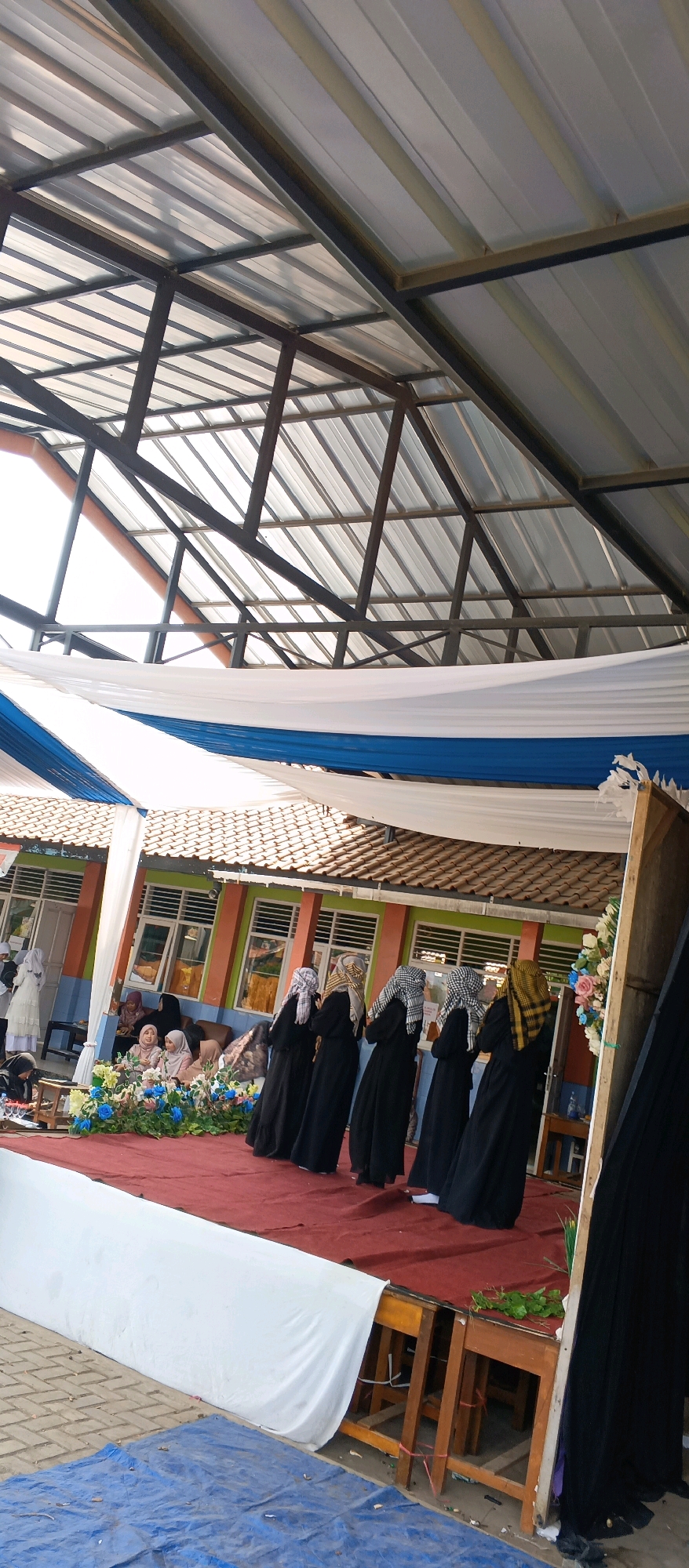 This image depicts an outdoor event taking place on a raised stage covered with a red carpet, likely in Palir, Indonesia. A group of six individuals, dressed in black robes and headscarves (hijabs), are standing on the stage, facing away from the camera and towards a building. The building has a tiled roof and colorful walls, with windows that appear to have white blinds. The stage is decorated with blue and white draped fabric overhead, and a floral arrangement with blue and white flowers is visible on the left side of the stage. In front of the stage, a blue tarp covers a section of the ground, which appears to be paved with bricks or tiles. The scene is set under a metal roof structure with translucent panels, suggesting it might be an open-air pavilion or a covered outdoor space.