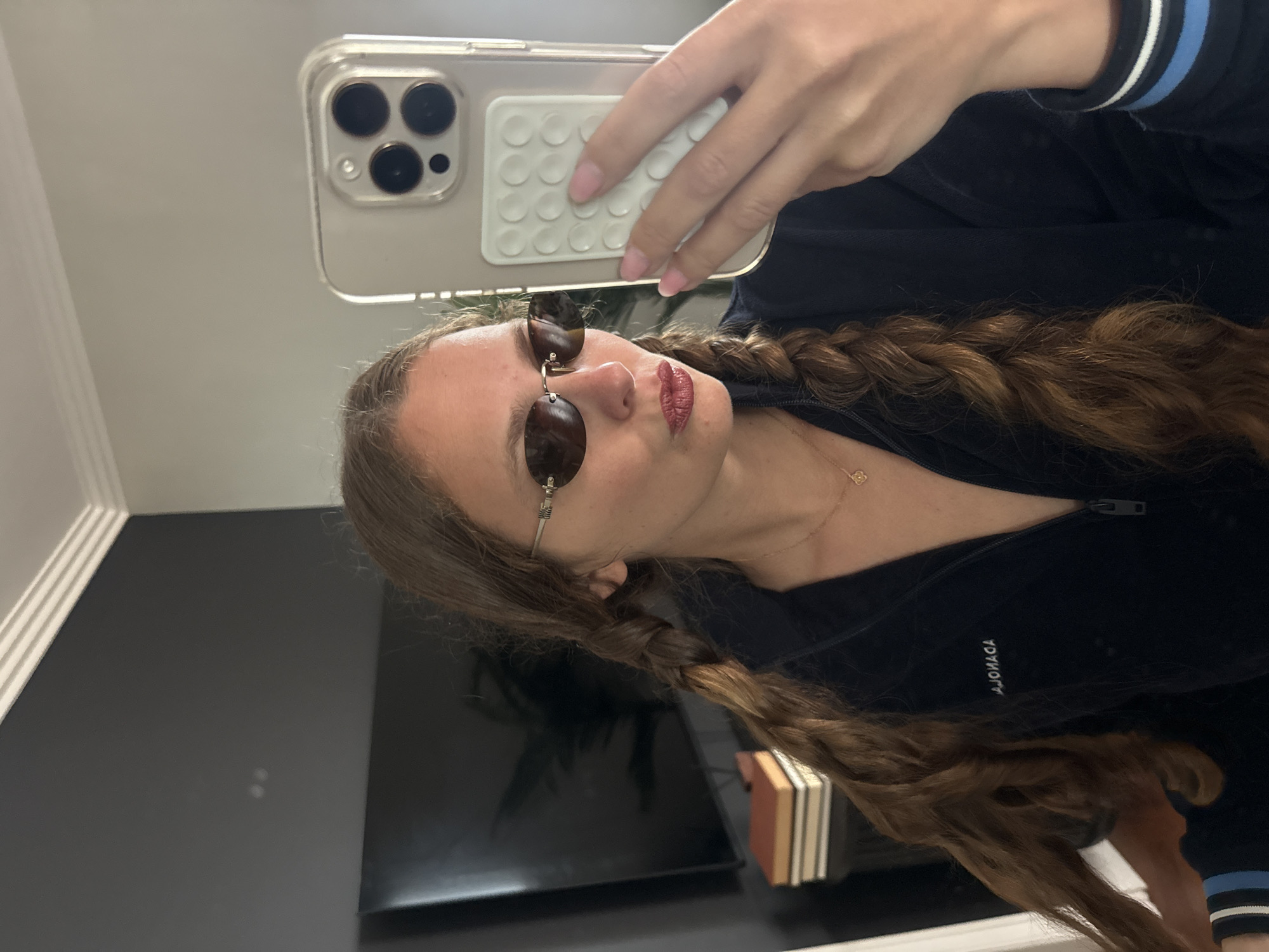 A young woman takes a selfie in a room with a reflective dark surface, possibly a table or counter. She is wearing sunglasses with oval, tortoise-shell-patterned lenses, a dark-colored zip-up jacket with blue and white striped cuffs, and a delicate gold necklace. Her long, light brown hair is styled in two braids. She holds an iPhone with a clear case and a white, circular-patterned accessory attached to the back. The background includes a dark gray wall with white trim. The location cannot be determined to be in Cape Town, South Africa.