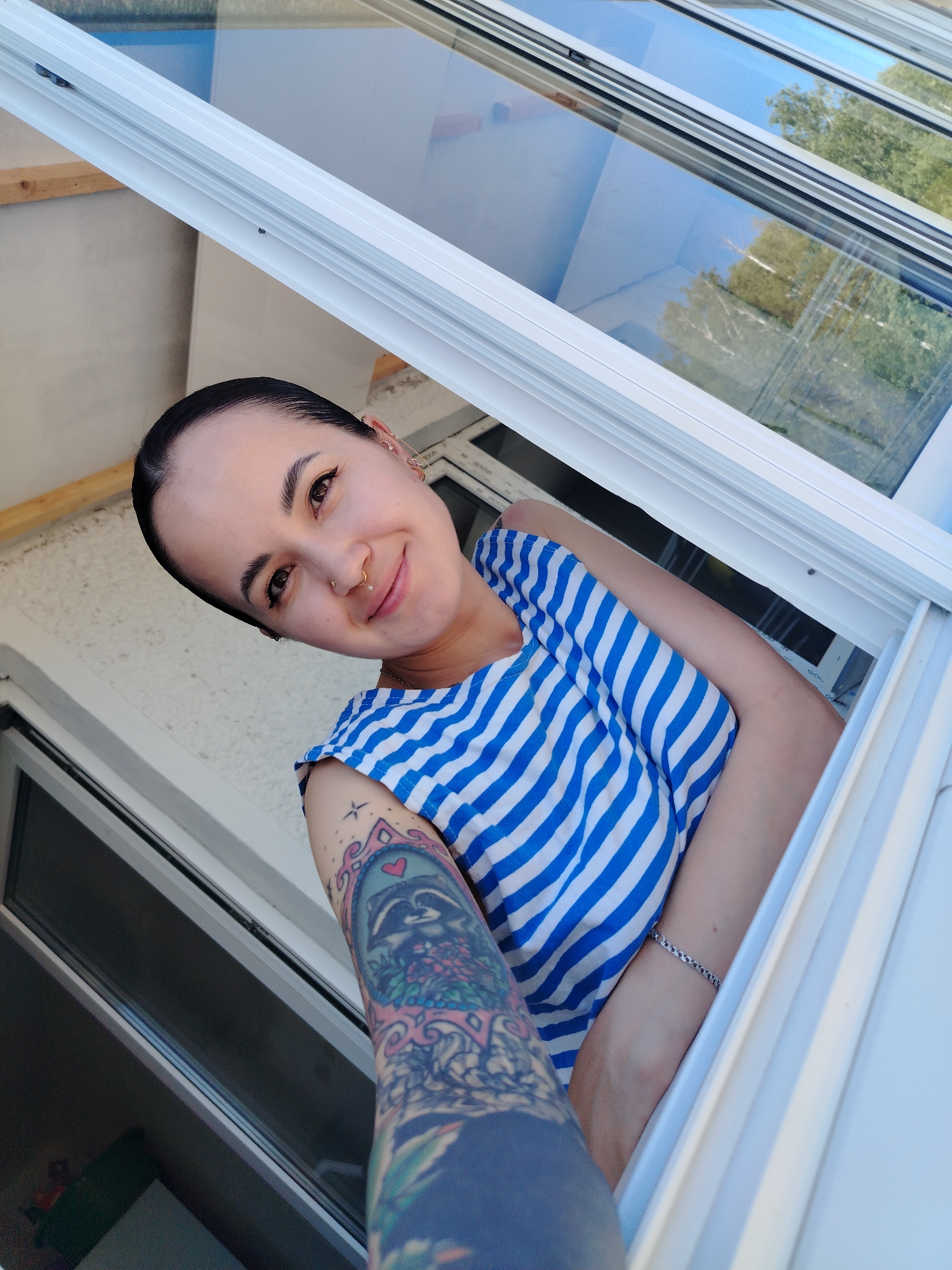 A young woman is leaning out of an open window. She has dark hair pulled back, light skin, and a nose ring. She is wearing a blue and white striped top. Her left arm is visible, covered in a colorful tattoo. A silver bracelet is on her right wrist. The window frame is white. Through the window, trees and the sky are visible. The location is Noviy, Russia.