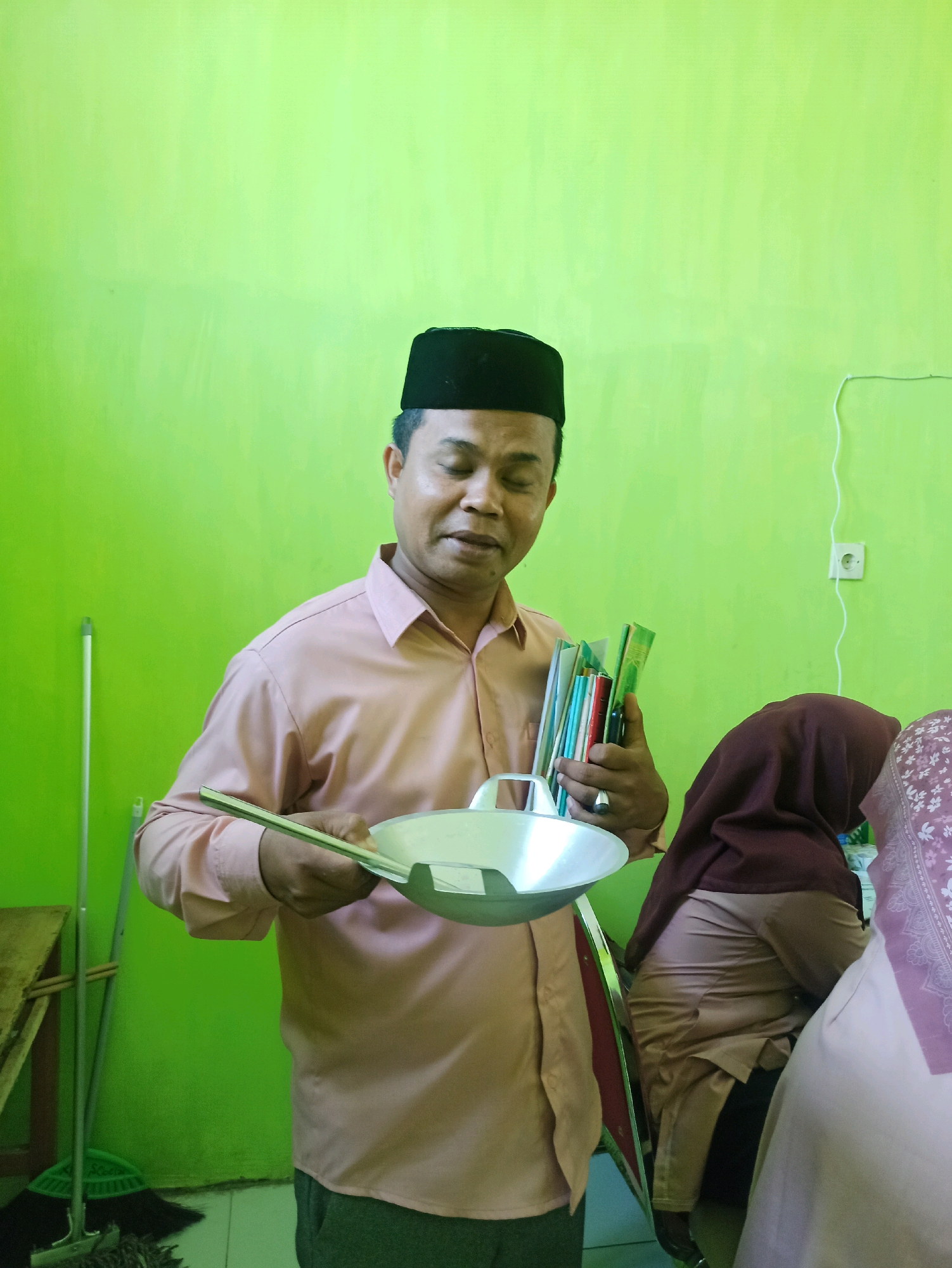 A man wearing a peach collared shirt and a black peci hat stands in front of a bright green wall, holding a frying pan and a stack of books. He is looking down and to his left. To his right, the heads of two individuals wearing hijabs are visible, also looking downwards. In the background, a light switch is visible on the wall. To the left of the man, a broom and a portion of a wooden table can be seen. The man is in the city of Panggungan, Indonesia.