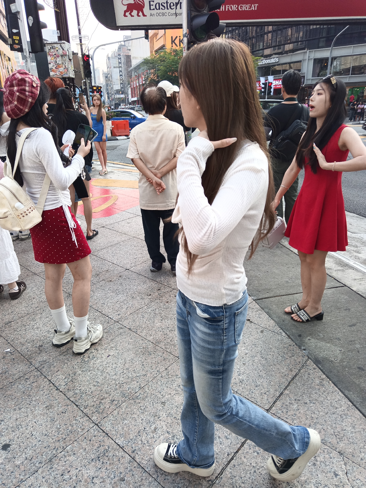 A street scene in Kuala Lumpur, Malaysia, shows several people walking along a busy sidewalk. In the foreground, a woman with long brown hair, wearing a white long-sleeved top, blue jeans, and black and white platform sneakers, is walking away from the camera, her left leg extended forward. To her left, another woman in a red dress and black slide sandals is facing away, her right hand raised. Behind her, a woman in a red and white checkered beret, white long-sleeved top, and red polka-dot skirt, with white socks and sneakers, is holding a phone in her left hand and a white portable fan in her right. She is looking at the woman in the blue dress. The woman in the blue dress is standing near a traffic light and holding a phone, taking a selfie. In the background, several other individuals are visible, along with buildings, traffic lights, and street signs, including one that reads "Eastern An OCBC Company". The ground is paved with granite tiles, and a yellow and red painted pedestrian crossing is visible in the mid-ground.