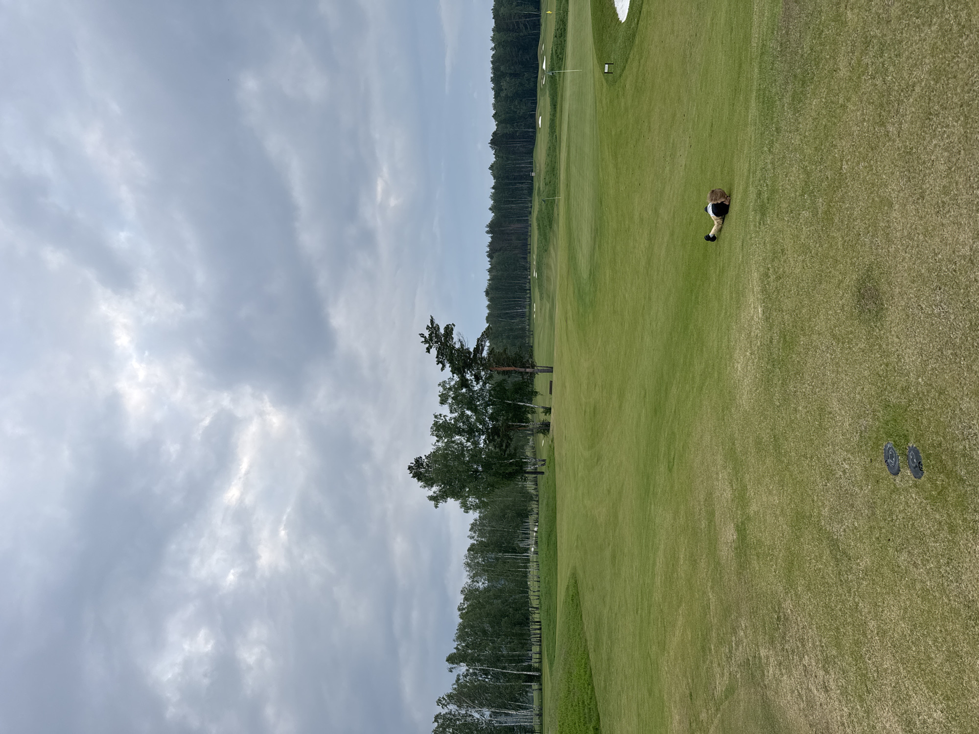 A person is lying face down on a golf course. The person is wearing dark shorts and a light-colored shirt with dark stripes. The person is on the right side of the frame, in the middle distance. The golf course is green with manicured grass and some bare patches. In the background, there is a line of trees, and beyond that, more trees and a cloudy sky. There are several sand traps visible on the course, as well as flagsticks. The location is not specified, but the context suggests a golf course in Krasnoyarsk, Russia, given the user's prompt. The overall scene is one of a person in a prone position on a golf course, under an overcast sky.