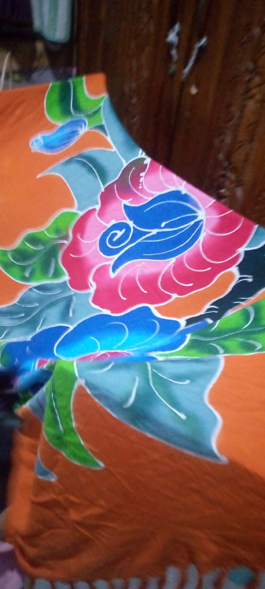 The image is a close-up, low-angle shot of a colorful batik fabric with floral patterns. The fabric is predominantly orange, with large stylized flowers in shades of pink, red, and blue. Green and teal leaves with white outlines surround the flowers. The batik is draped over an unseen object, casting shadows and creating folds. In the blurred background, a dark wooden surface, possibly furniture, is visible. The scene is indoors, but the specific location in Sumberwono, Indonesia cannot be determined from the image. There are no people or visible actions.