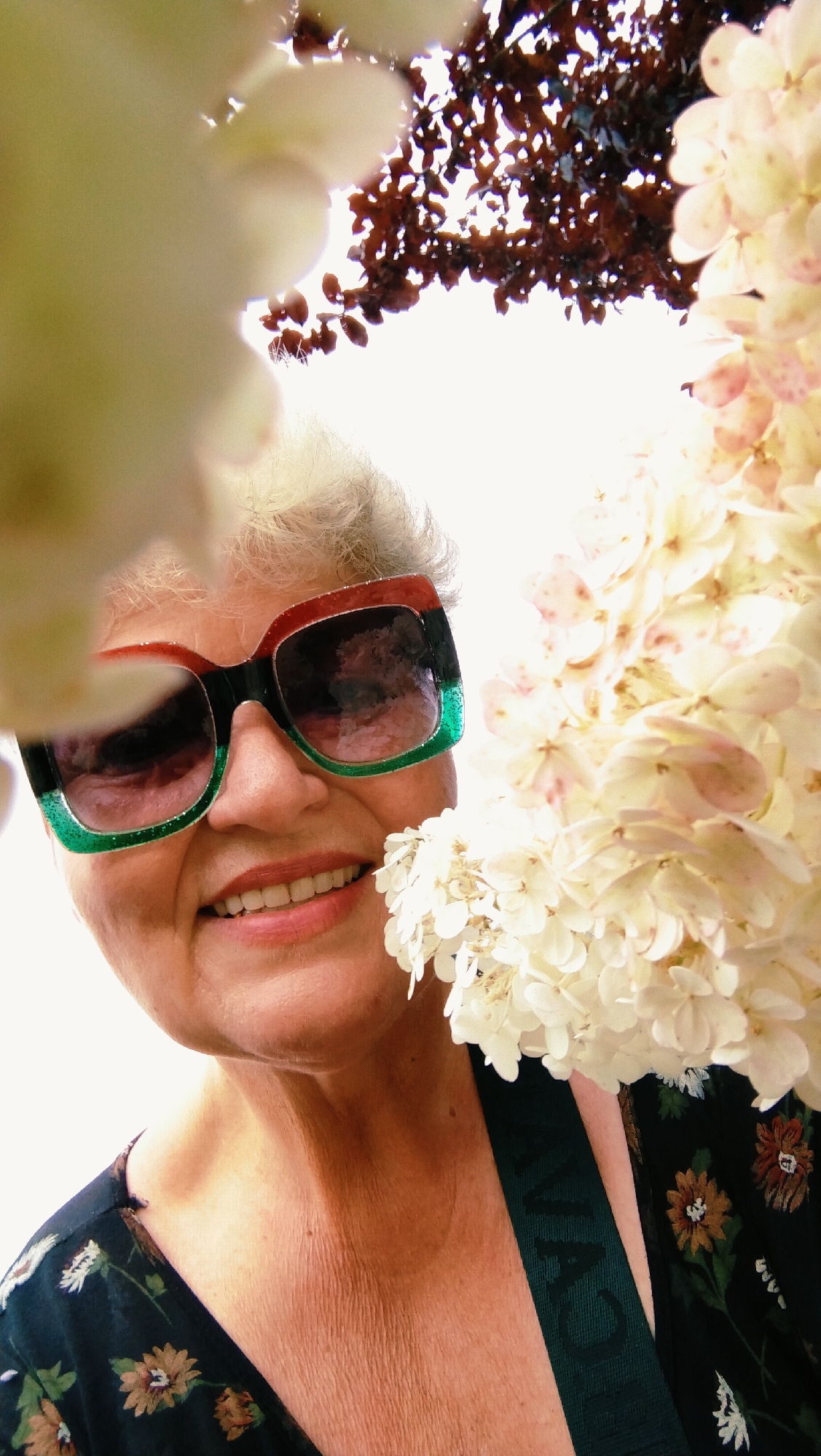 A close-up shot captures a woman with short, grey hair smiling as she peers through the blossoms of a flower. She is wearing large, rectangular sunglasses with frames that are black, red, and green. Her lips are parted, revealing her teeth. The woman is wearing a dark floral-patterned dress. The background is bright, with hints of brown leaves and more white blossoms. The image is taken from a low angle, looking up at the woman. There is no identifiable information about the city of Horishni Plavni, Ukraine, within the image.