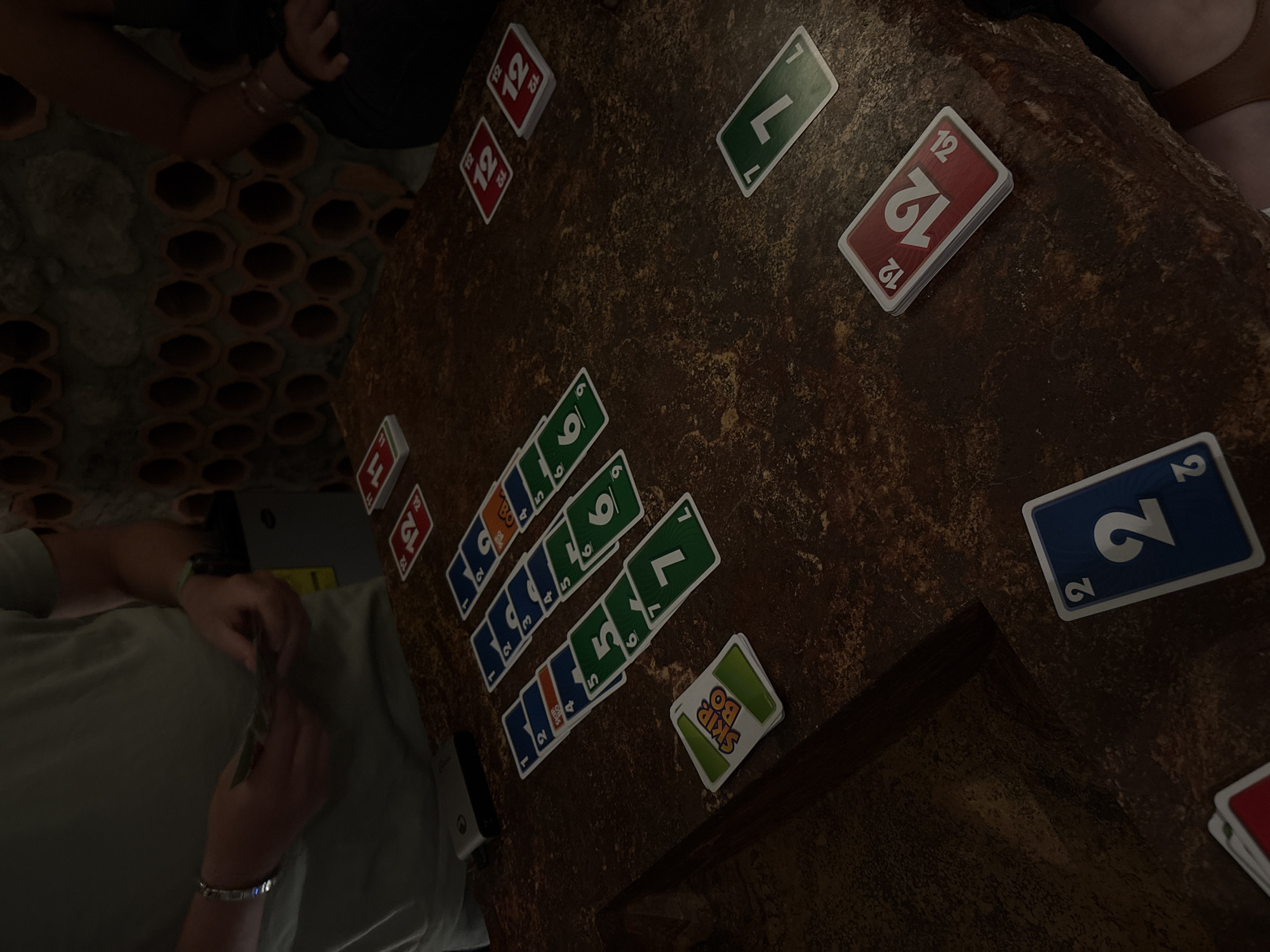 The image depicts a Skip-Bo card game in progress on a dark, textured surface resembling a wooden table. A person with a light-colored shirt is holding cards, suggesting they are a player in the game. Several stacks of Skip-Bo cards are arranged in a grid-like pattern, including stacks with visible numbers such as 2, 4, 5, 6, 7, 11, and 12. Other cards are scattered around the table. The setting appears to be indoors, with decorative elements such as rows of small terracotta pots visible in the background. The image was taken in San Bernardino Tlaxcalancingo, Mexico.