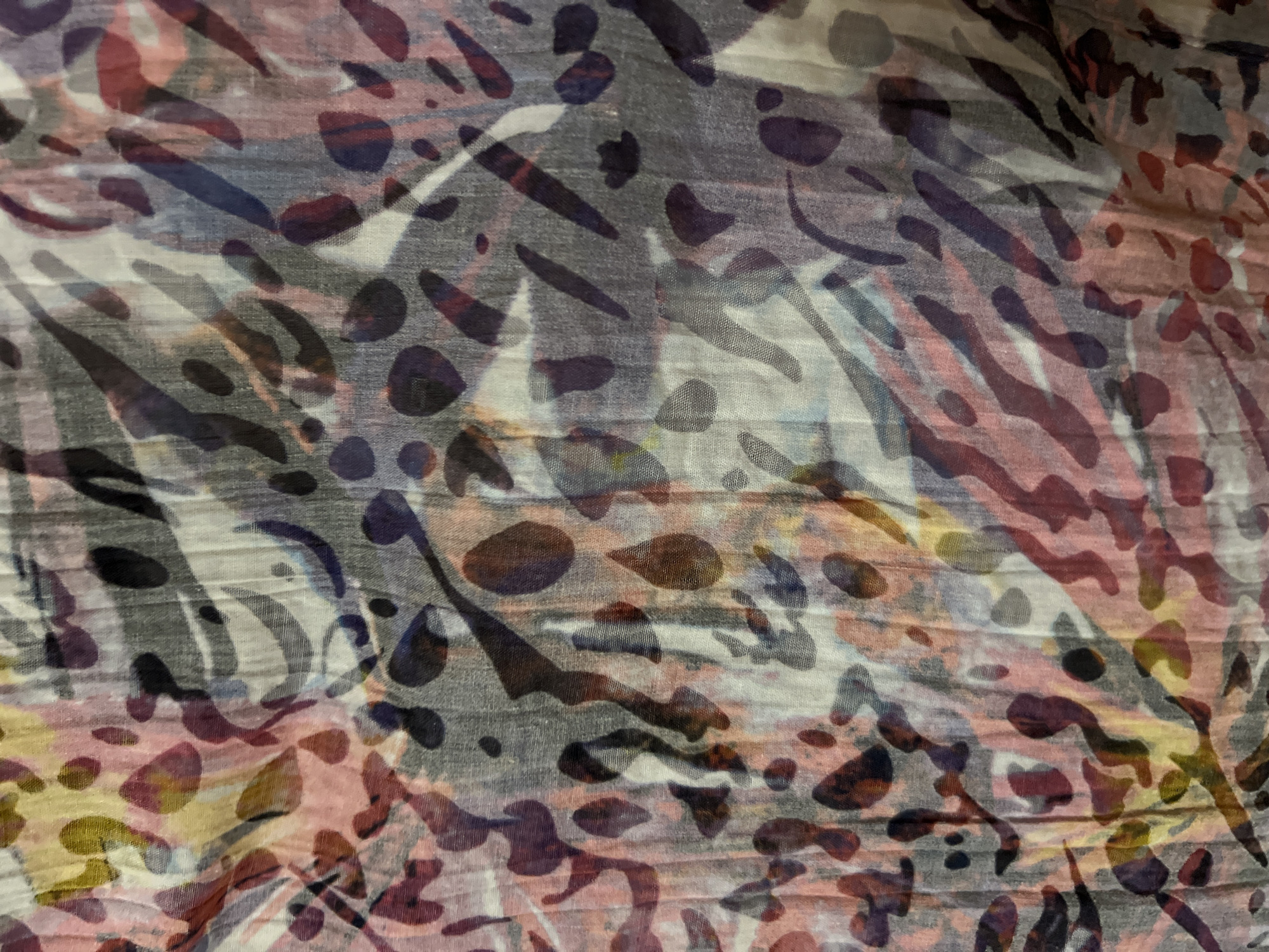 The image displays a close-up view of a fabric with an abstract, watercolor-like pattern. The pattern features a mix of organic shapes and blotches in shades of pink, purple, yellow, gray, and black. The fabric appears to be thin and slightly creased, suggesting it could be a scarf, blouse, or other lightweight garment. The lighting casts subtle shadows and highlights, emphasizing the texture of the material. There is no visible indication of the city of Minsk, Belarus, or any specific location. The image is a detail shot of fabric and does not contain any people, actions, interactions, or environmental context.
