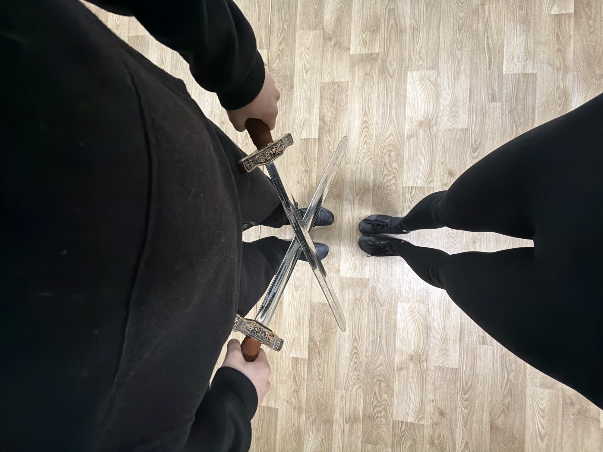 A high-angle shot shows two individuals facing each other on a light wooden floor, possibly laminate. One individual, on the left, is wearing dark, loose-fitting clothing with long sleeves. They hold a medieval-style sword with a silver blade and a brown handle, displaying intricate detailing. The sword's point rests on the floor. The other individual, on the right, is wearing dark leggings and black shoes. Their posture suggests they are also holding a sword, with the blade and hilt visible but partially obscured by the first sword. The swords intersect near the center of the frame. The location is unknown and there is nothing to indicate it is the city of Plovdiv, Bulgaria.