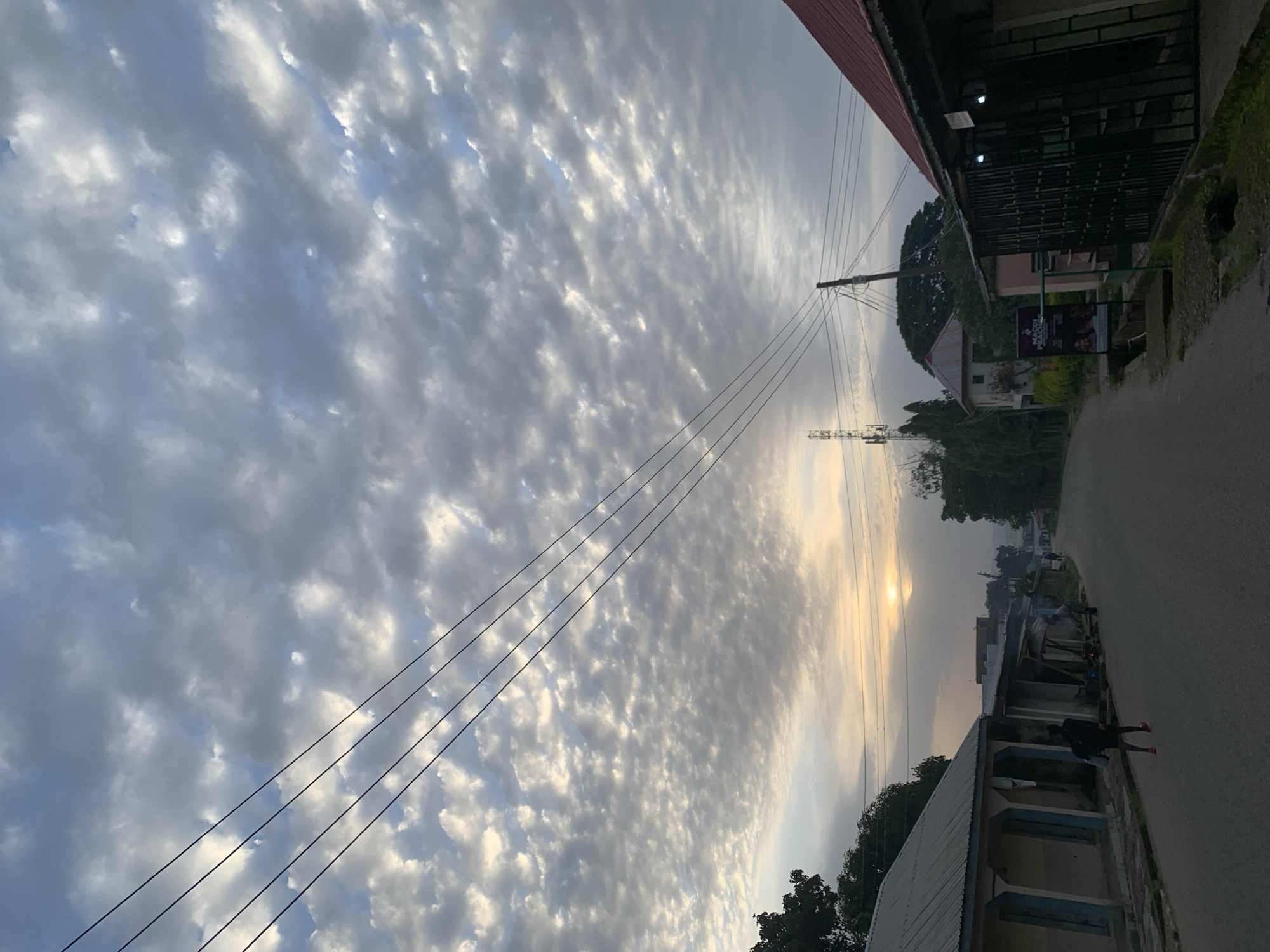 The image depicts a street scene in Jos, Nigeria, at what appears to be early morning or late evening, suggested by the soft sunlight breaking through a heavily clouded sky. The sky is filled with a textured pattern of clouds, with a faint glow of the sun visible near the horizon, partially obscured by the clouds. Several power lines are strung across the upper portion of the frame, contributing to a sense of urban infrastructure.
The street itself is paved and curves gently into the distance, lined by buildings on both sides. On the left, a single-story building with a corrugated metal roof and light-colored walls is visible, featuring a shaded porch area with columns. Further down the street, more buildings with similar roofing materials are present, some appearing to be shops or residences. One building on the right has a prominent red metal roof and a fenced-off storefront.
Two individuals are visible on the street. One person, wearing dark clothing and red shoes, walks away from the camera down the street. Another figure, also in dark clothing, is seen further in the distance near the buildings on the left.
In the background, trees and vegetation are present, interspersed with more buildings and a cell tower, indicating a mix of natural and developed environments. The overall atmosphere is quiet and subdued, with the cloudy sky and soft light creating a somewhat muted visual palette.