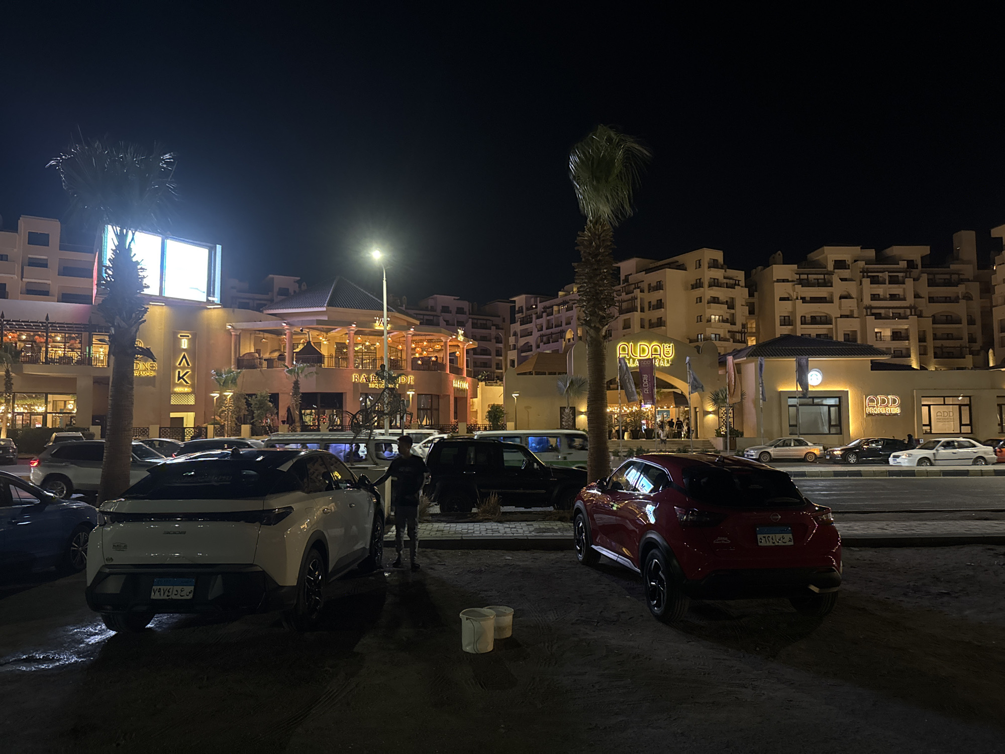 Here is a detailed description of the image:
The image, taken at night, showcases an outdoor scene with several buildings, cars, and people. The buildings, tan in color, are illuminated with lights and appear to be either restaurants or businesses. There are also some palm trees, typical of a tropical or subtropical environment.
In the foreground, there are several cars parked in what looks like a sandy or dirt parking lot. There are two cars that are prominently visible: a white SUV and a red SUV. A person is standing near the white SUV. There are also two buckets on the ground between the cars, suggesting that someone might be washing or cleaning the vehicles.
Behind the parked cars is a road where other vehicles are parked. Beyond the road are the buildings. The lighting in the scene is a mix of ambient light from the buildings and street lights.
The license plates on the vehicles indicate that they are from Egypt.
