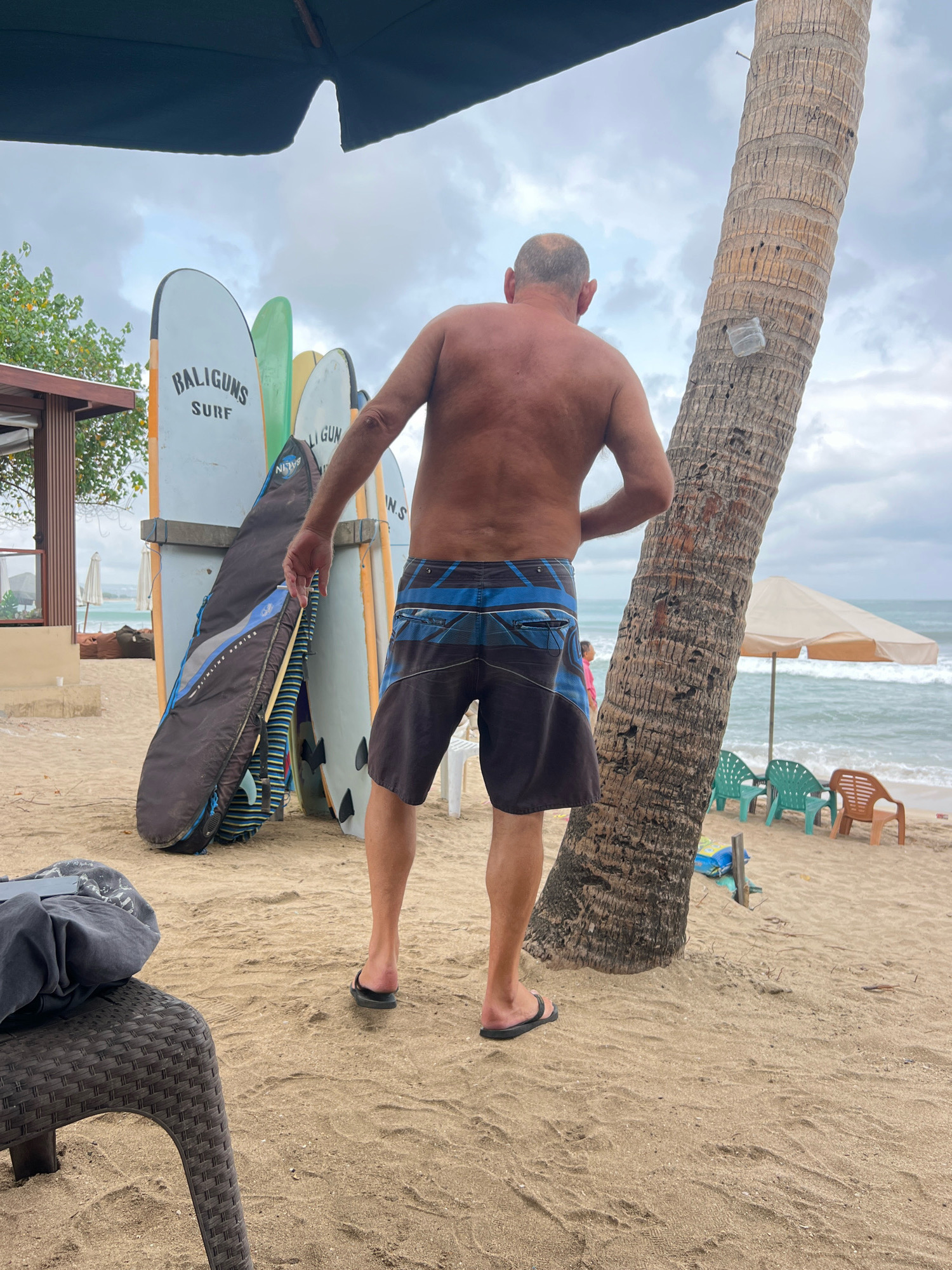 A man, seen from behind, stands on a sandy beach in Kuta, Indonesia, near a cluster of surfboards. He is wearing dark swim shorts with blue stripes and black flip-flops. Behind him, several surfboards are propped up, some with the branding "BALIGUNS SURF" visible. A large palm tree trunk stands to his right. The background shows the ocean with moderate waves, a beach umbrella, and a few empty plastic chairs. The sky is overcast, suggesting cloudy weather. In the foreground, a dark gray bag or piece of clothing rests on a wicker chair.