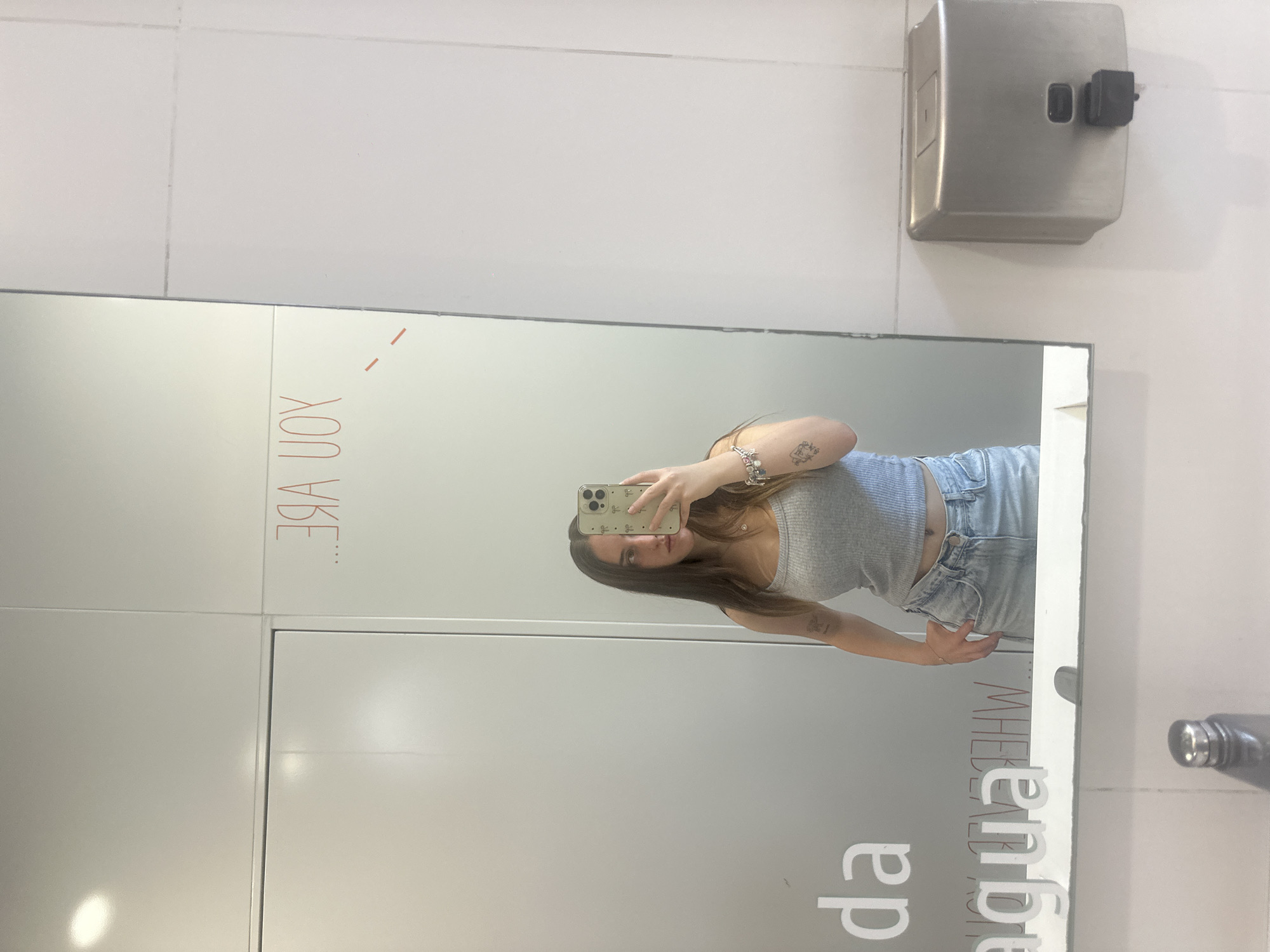 A young woman is depicted taking a mirror selfie in what appears to be a public restroom or changing room. The image is rotated 90 degrees counter-clockwise, showing the woman horizontally. She is wearing a grey ribbed crop top and light-wash blue jeans. Her left arm, adorned with bracelets and a small tattoo, holds a smartphone with a light green patterned case, reflecting her face and upper body. Her expression is neutral.
The background consists of plain white/light grey tiled walls and a large mirror. A metallic wall-mounted dispenser, possibly for soap or paper towels, is visible in the upper right corner. In the lower right, the cap of a water bottle or thermos is partially visible.
Visible text on the mirror appears backward due to reflection. In the upper left, red text reads "... YOU VBE..." (likely "YOU VIBE..."). In the lower right, partially obscured text in what seems to be white or light grey font reads "...DA AGUA MHE...", possibly "da água" which means "from the water" in Portuguese or Spanish.