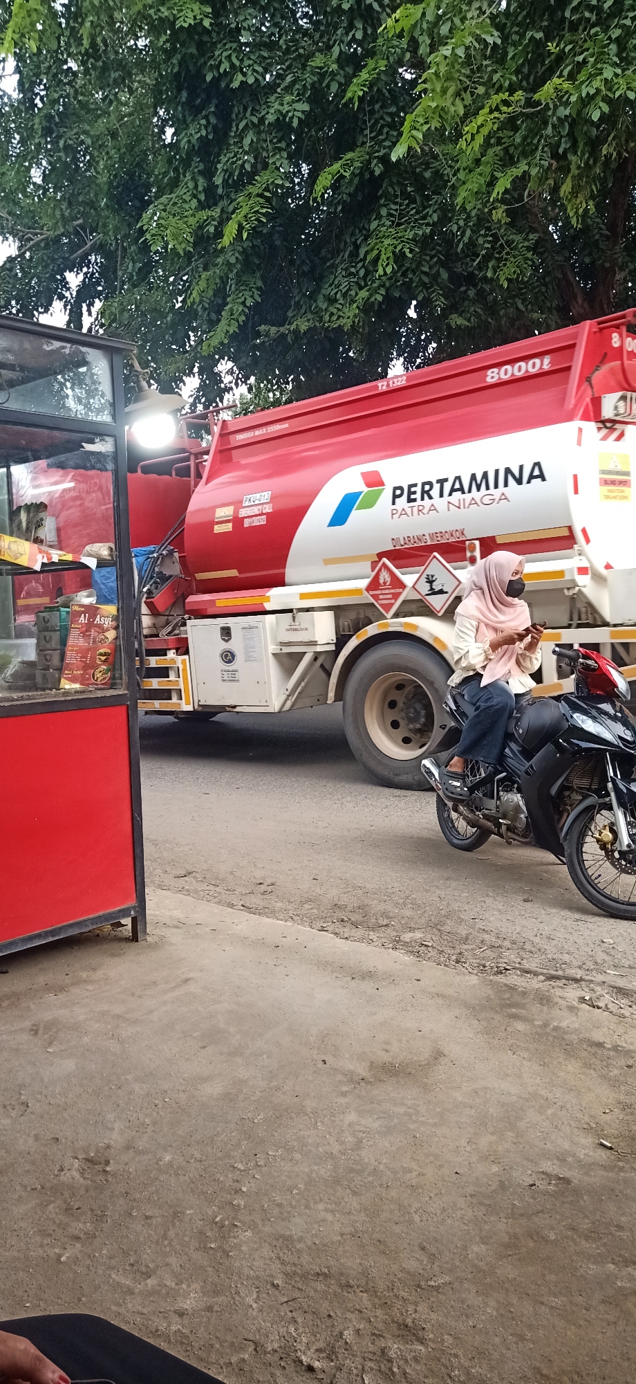 A red and white Pertamina Patra Niaga fuel tanker truck is parked on a street in Gampongteungoh, Indonesia. The truck has "8000ℓ" printed on its side, along with the Pertamina logo. A woman wearing a light pink hijab and a dark mask is sitting on a black motorcycle next to the truck, looking at her phone. She is wearing a light-colored blouse and dark pants. The scene appears to be during the daytime, with trees in the background. To the left of the truck is a red food stall with a glass display case, showcasing some food items and a menu board. The menu board displays text that includes "Al-Asyi" and images of food. The ground is paved and shows signs of wear. The overall atmosphere seems to be a typical street scene in a town.