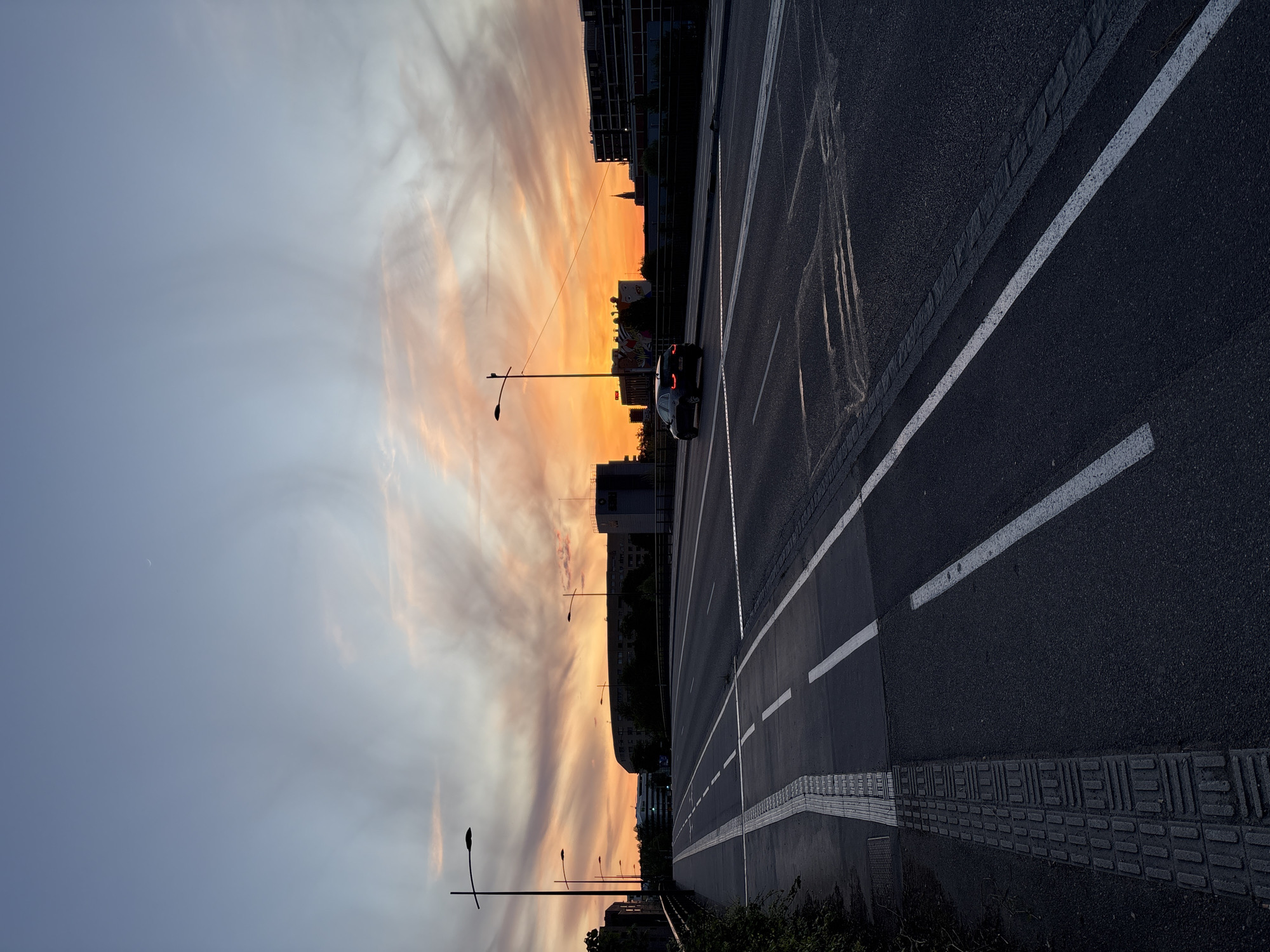 The image depicts an urban sunset scene from Strasbourg, France. The perspective is a low-angle shot along a road, presumably a bridge or elevated highway, towards the west. The sky exhibits a vibrant orange and pink gradient, transitioning to lighter hues near the horizon, indicating sunset. Wispy clouds are visible across the sky. Streetlights are sparsely placed along the roadside. A car with illuminated taillights is visible on the road.
The road itself is dark asphalt with white lane markings. A textured tactile paving can be seen. Buildings and trees are silhouetted against the sunset, creating a distinct contrast. The sky is partially obscured by buildings. The overall tone is peaceful, illuminated by the warmth of the setting sun. The crescent moon is also visible.
