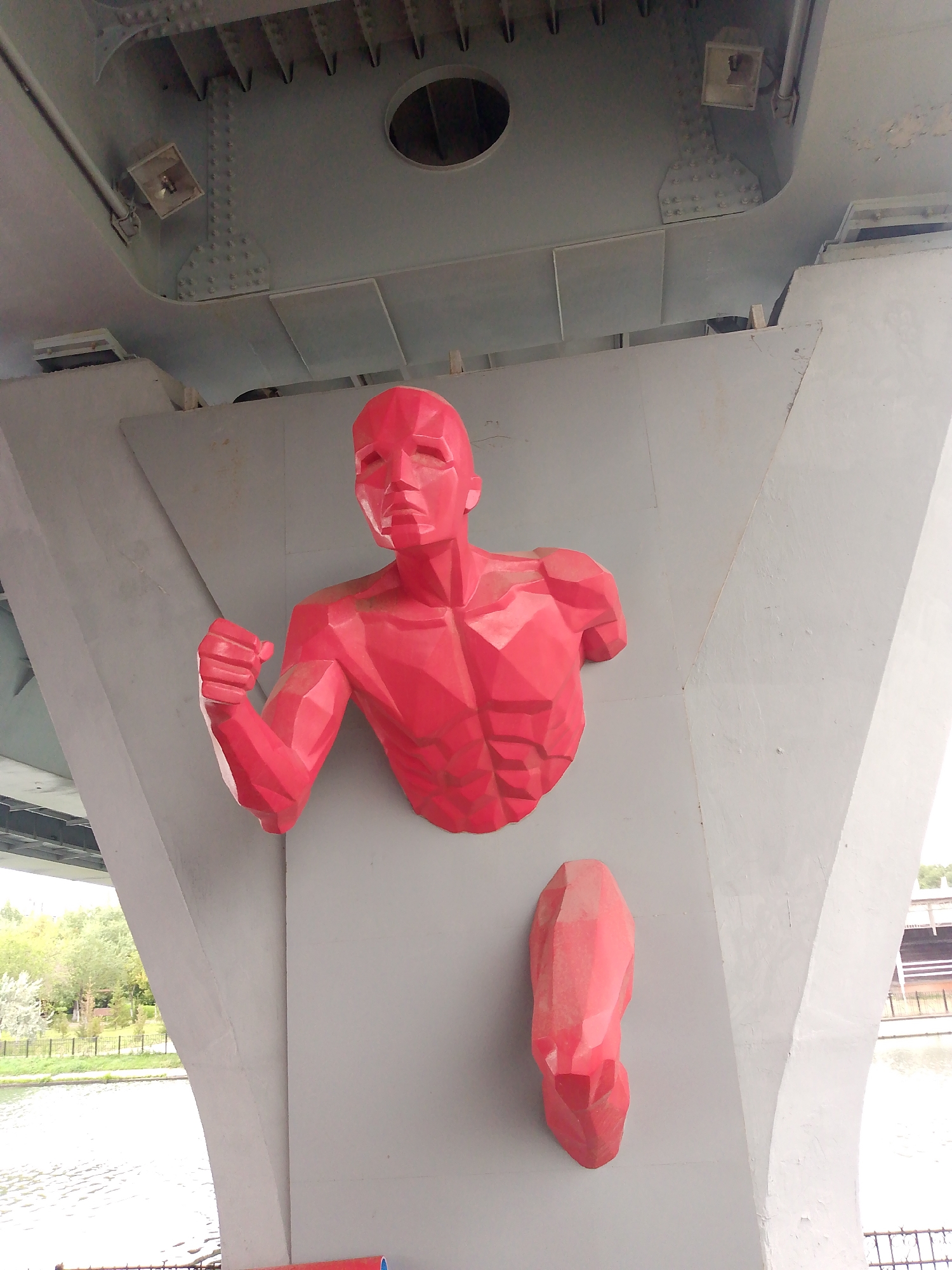 The image shows a large, stylized, red sculpture of a human torso and a detached leg attached to the underside of a concrete structure, possibly a bridge. The sculpture is made of geometric planes and is positioned against a gray concrete wall. The torso is depicted in a running pose, with one arm raised and clenched into a fist. The detached leg is positioned below the torso. In the background, there is a body of water, trees, and a railing. The overall scene suggests an outdoor urban environment. This sculpture is located on the bridge over the Ishim River in Astana, Kazakhstan.