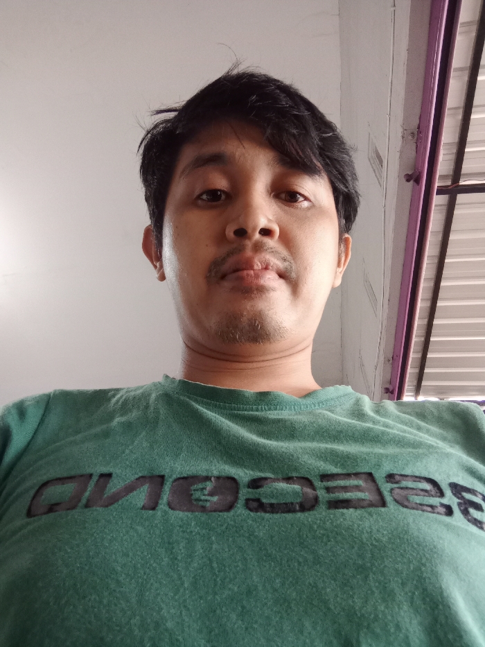 A medium shot displays a man looking directly at the camera from a low angle. He has dark hair, a mustache, and wears a green t-shirt with the word "SECOND" printed in black letters. The background reveals a white wall and a glimpse of what appears to be a building exterior, possibly a metallic roof or paneling, accented with a purple frame.
