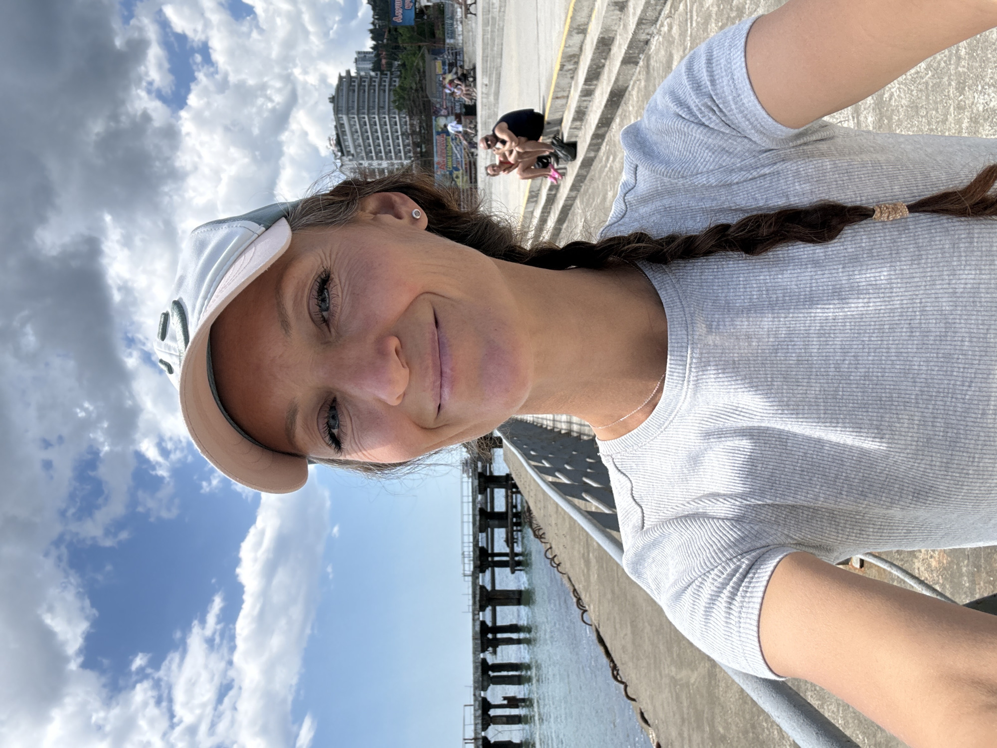 The image is a selfie taken outdoors on a sunny day with scattered clouds. The subject is a woman wearing a light-colored baseball cap and a light gray ribbed t-shirt. She has a braid of dark hair down her back and a small silver earring in her visible ear. Her face is turned towards the camera, and she is smiling.
In the background, to the left, is a body of water with a pier extending out from the left edge of the frame. The pier is supported by concrete pillars and appears to be weathered. To the right of the woman, there are concrete stairs leading up to a paved area. On this paved area, several people are visible, some sitting on the stairs or benches. Further in the background, on a hill, are buildings, suggesting an urban or resort area. The overall setting appears to be a waterfront promenade or embankment. The text on a distant sign includes Cyrillic script, and based on visual cues like the architecture and landscape, the location is likely Alushta, Ukraine, a city on the Crimean Peninsula known for its seaside resorts.