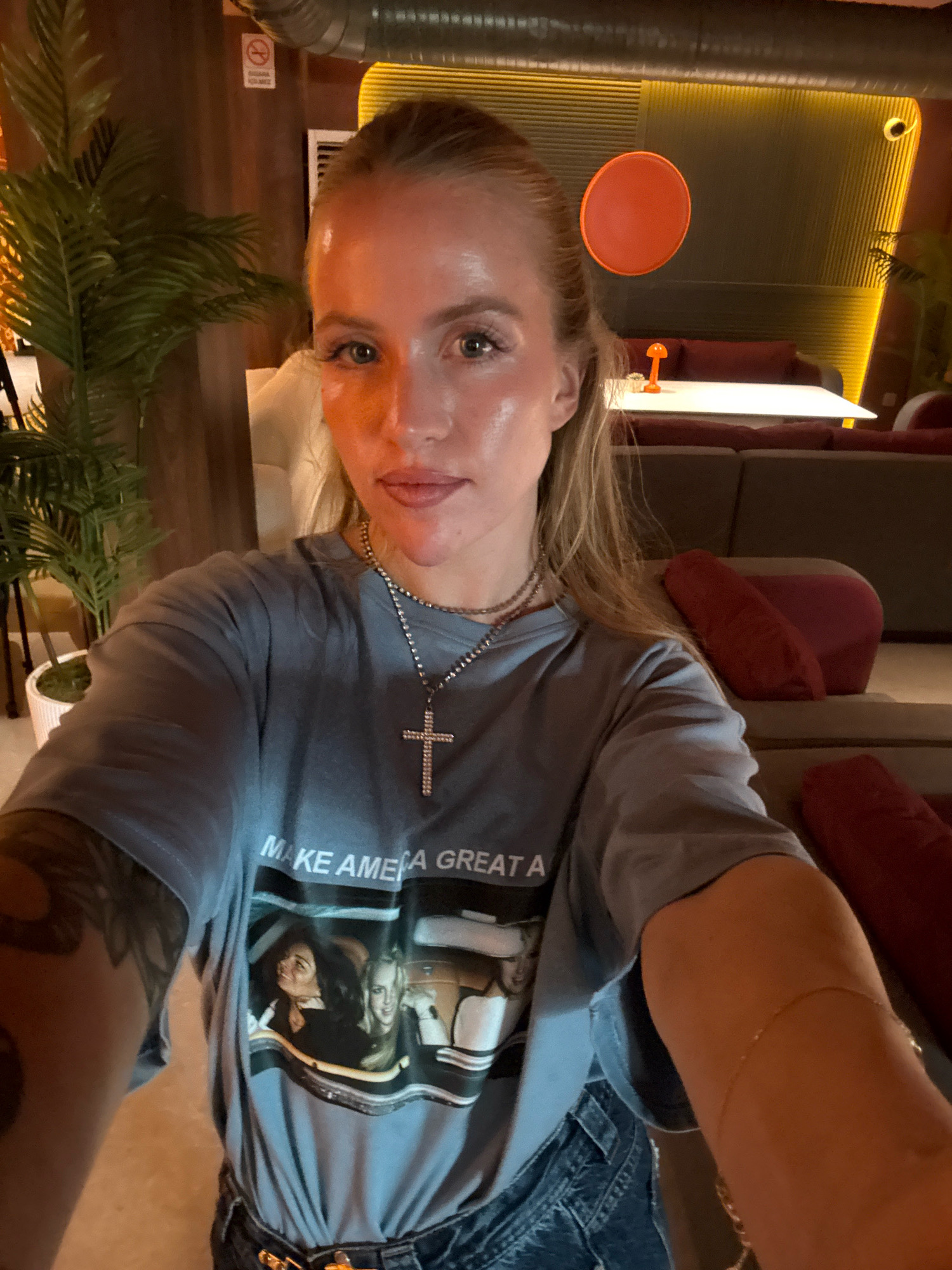 A young woman with blonde hair pulled back in a ponytail takes a selfie in a dimly lit, modern indoor space, possibly a restaurant or lounge. She is wearing a light blue t-shirt with a graphic that reads "MAKE AMERICA GREAT A" and features an image of two women in a car. She also wears a silver cross necklace and a gold bracelet on her right arm. The background shows a modern seating area with dark sofas and red pillows, along with some potted plants and decorative lighting. A sign with a red circle and a diagonal line, typical of a "no smoking" sign, is visible on a wooden pillar in the upper left. The overall ambiance is warm and inviting, with a mix of natural and artificial lighting.