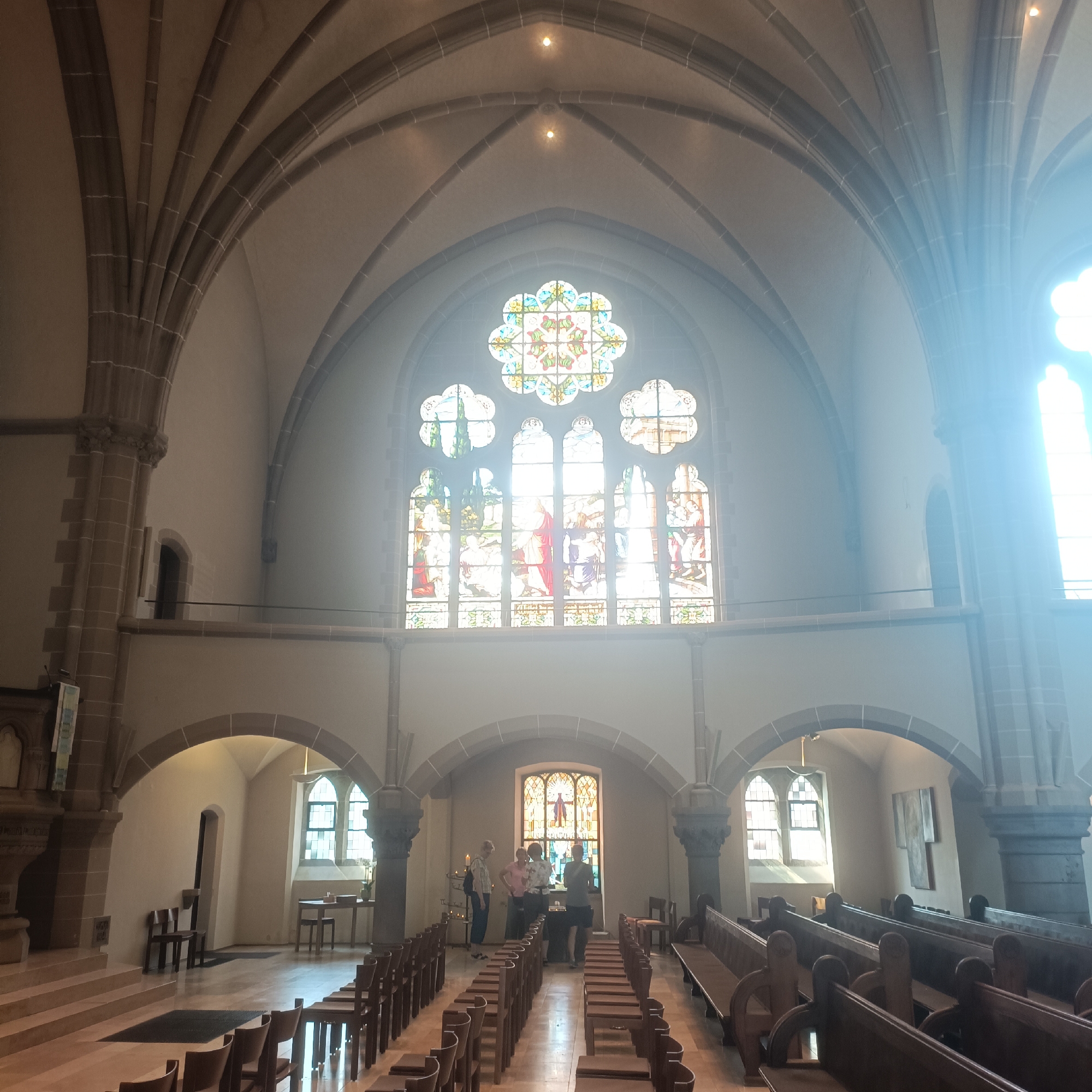 This image shows the interior of a church, likely St. Georg's Church in Bad Nauheim, Germany, given the architectural style and stained glass windows. The scene depicts a nave with rows of wooden pews leading towards an altar area. Above the altar is a large, ornate stained glass window featuring a central circular motif and surrounding rectangular panels. Sunlight streams through this window, creating a bright, hazy effect. In the foreground, several people are gathered near a table in front of the altar, appearing to be in conversation or examining something. The architecture features high vaulted ceilings with stone ribs, arched doorways, and a decorative stone column on the right. The overall impression is one of solemnity and grandeur, typical of a historic church interior.
