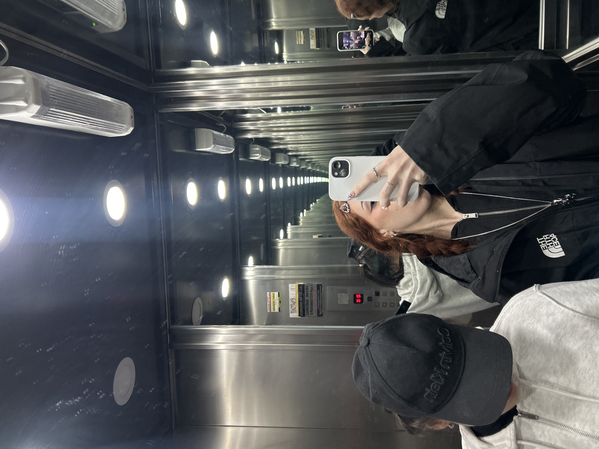A medium, angled shot captures a woman taking a selfie in an elevator, reflected in the elevator's mirrored walls and ceiling. The elevator's interior is metallic, with rows of illuminated, round ceiling lights stretching into the reflected distance. The woman is holding a white smartphone.
She has reddish-brown hair and is wearing a black jacket with a visible "The North Face" logo on the chest. On the elevator floor, partially visible, is another person with a black "Calvin Klein" baseball cap and a light-colored hoodie.
Elevator details are visible, including a floor indicator displaying "35", buttons, and an information sticker with "CAPACIDAD MAXIMA 3 PERSONAS (225 Kgs.)" written on it.