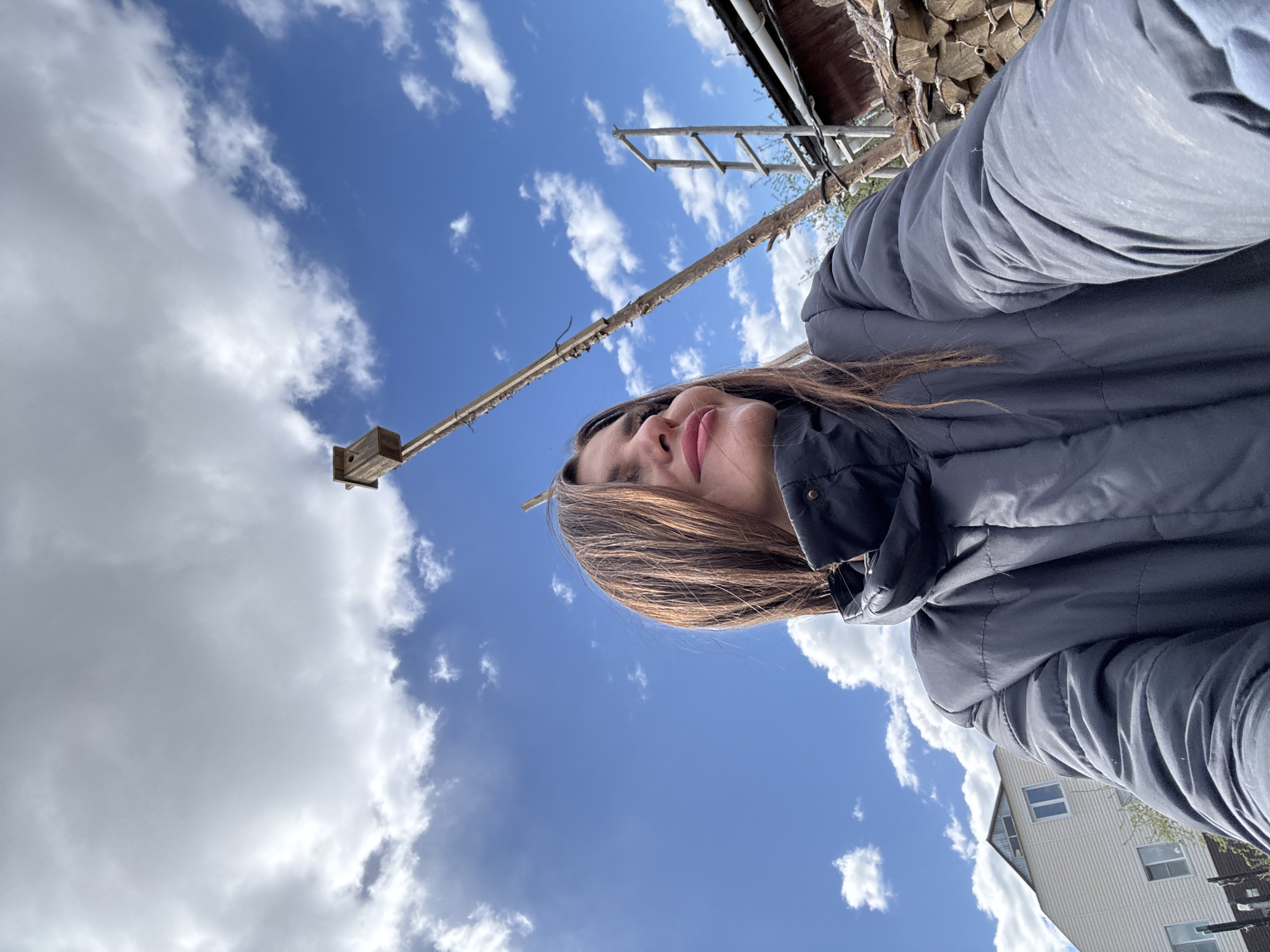 A woman in a black jacket is the central subject, appearing to be taking a selfie. The sky is visible with a mix of blue sky and white clouds. A wooden structure with a box on top extends into the sky. A wooden ladder and stacked wood are also visible near a building. The location appears to be in a residential area of Dubovaya, Russia, given the presence of houses in the background.