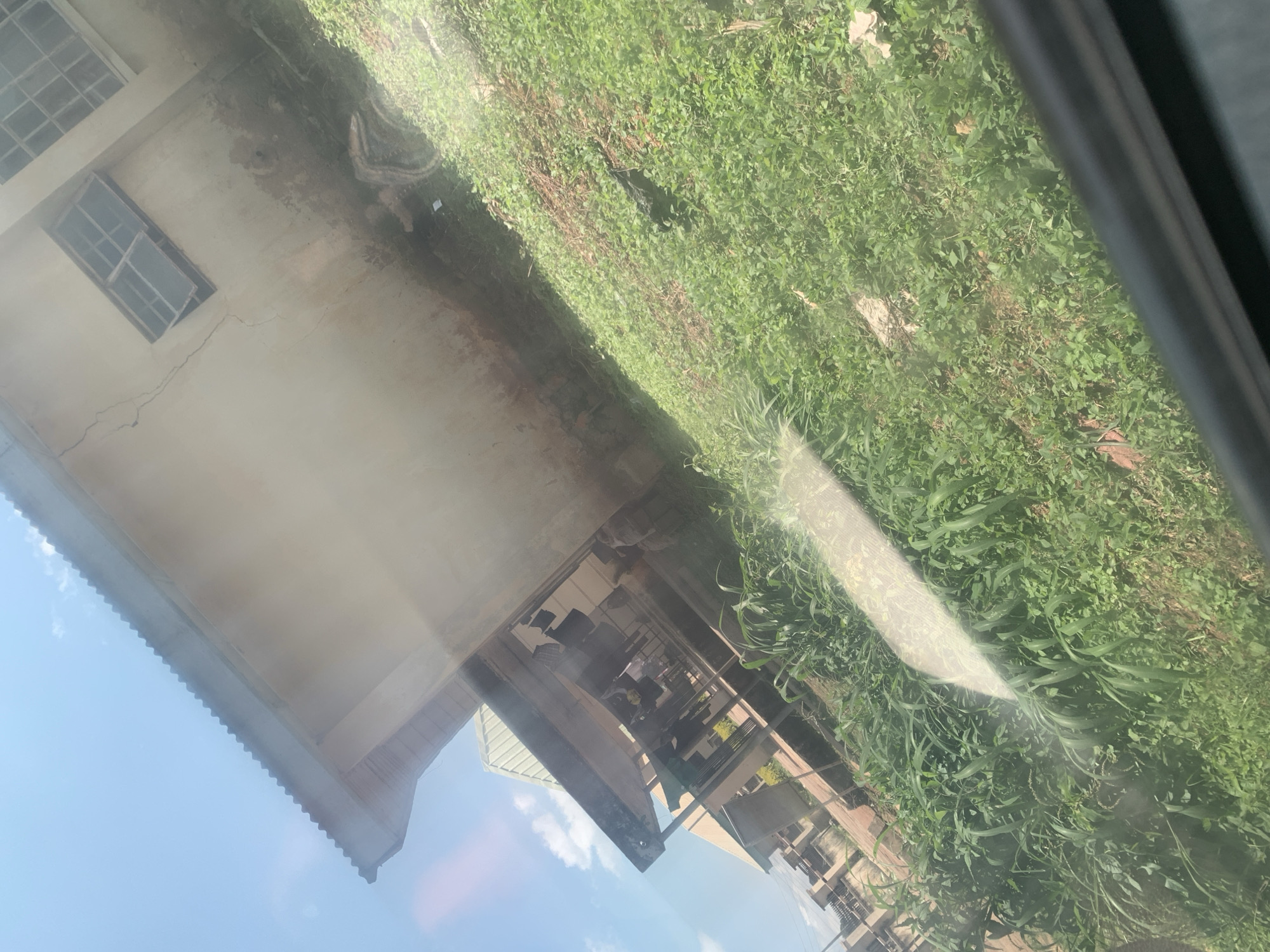 The image shows an outdoor scene, possibly in the city of Jos, Nigeria, with a building and surrounding vegetation. The building appears to be made of concrete and has a corrugated metal roof. There are several windows visible on the building, some of which are open. The building's exterior shows signs of age and weathering, with some cracks and discolored patches. In front of the building, there is an area covered in lush green grass and some taller plants. A covered walkway or veranda extends from the building, supported by columns. Clothes appear to be hanging on a line under this walkway. The overall scene suggests a slightly neglected but lived-in environment, potentially a residential or institutional building. The sky is blue with some scattered white clouds, indicating it is daytime. The image is taken from a slightly elevated angle, possibly from a vehicle, as indicated by a dark horizontal bar at the bottom of the frame and a diagonal reflection across the scene.