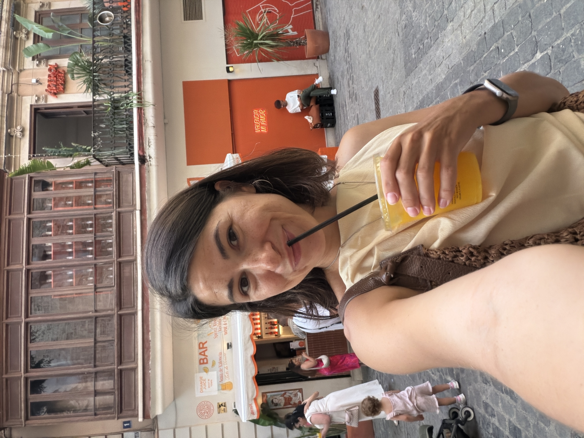 A woman in her early 30s is taking a selfie in an outdoor cafe setting. She is holding a yellow drink with a black straw and smiling at the camera. She is wearing a light beige sleeveless top and a brown bag over her shoulder. A man is seated on a bench in the background, and a child is standing next to a stroller. The cafe has an orange awning and an orange wall with neon signs. The overall impression is of a casual, sunny day in a European city.