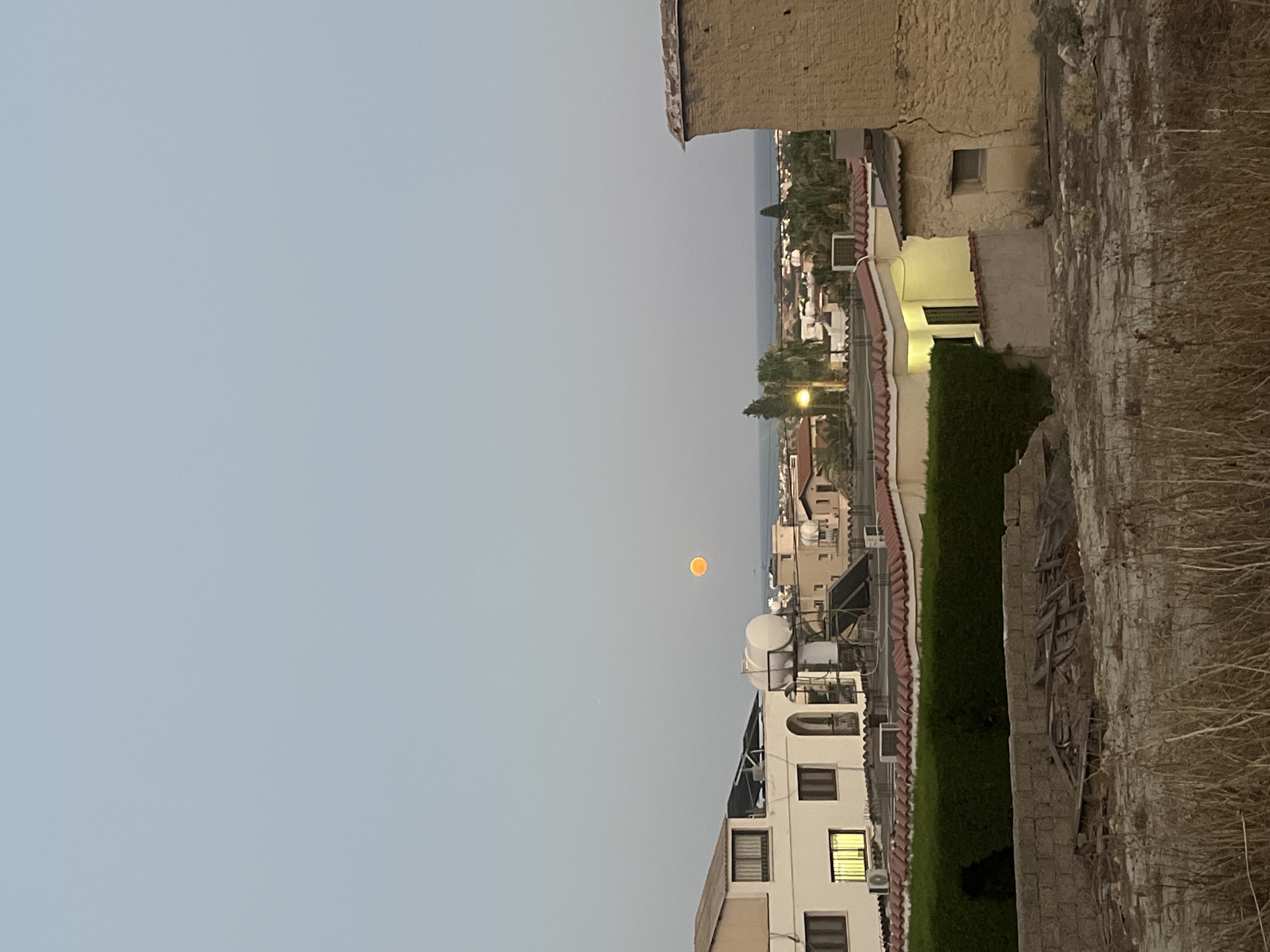 The image shows an aerial view of a residential area in Oroklini, Cyprus, at dusk. The sky is a pale blue, transitioning to a lighter hue near the horizon. A bright orange, circular object, likely the moon or sun, is visible in the upper-middle portion of the frame. Below the sky, a densely packed cluster of buildings with terracotta tiled roofs dominates the mid-ground. These buildings are of varying heights and architectural styles, some featuring white facades and balconies, others with tan-colored walls. Several solar water heaters are visible on some of the rooftops. A dark green hedge forms a boundary in front of some of the buildings. The foreground consists of dry, tall grass and a low stone wall, behind which are discarded wooden planks and other debris. In the distance, the blue expanse of the sea can be seen. The overall scene suggests a tranquil evening in a coastal town.