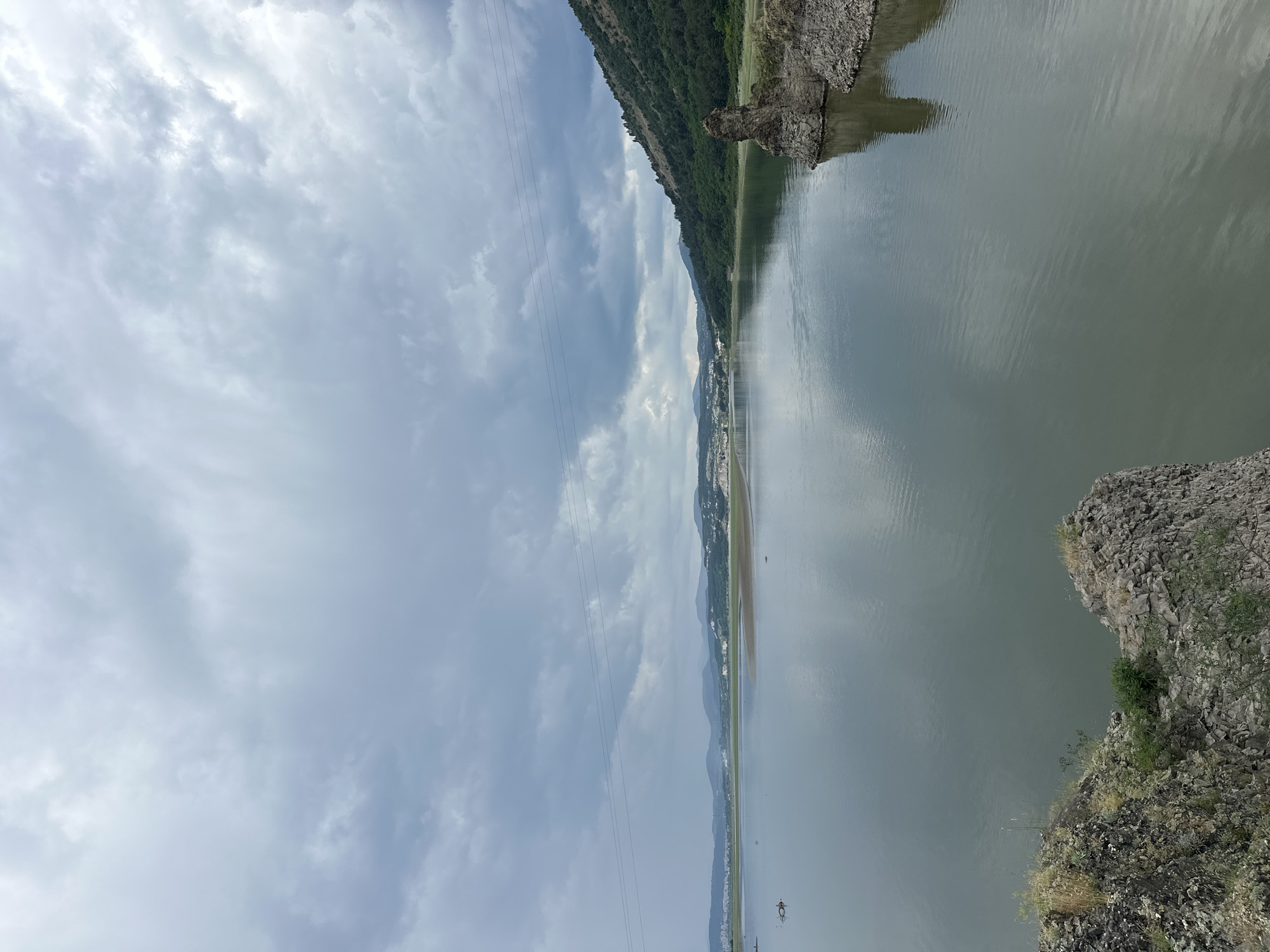 This image captures a wide, static landscape view of a large body of water, identified as the Kardzhali Reservoir near the village of Lisitsite, Bulgaria, under an overcast sky.
The upper approximately two-fifths of the frame is dominated by a heavily clouded, grey sky, exhibiting varying shades from light grey to darker, denser formations, with no visible sun or clear patches.
The central and lower portions of the image feature the expansive, calm surface of a reservoir, reflecting the muted tones of the sky. The water itself appears a deep, muted grey-green with subtle ripples visible across its surface, particularly closer to the viewer. On the far left side of the water, a dark, indistinct object, possibly debris or part of a submerged structure, is partially visible. Further out, small dark specks are scattered on the water, too distant for precise identification.
The foreground, at the bottom of the image, presents rocky terrain with sparse, dry vegetation, indicating the photograph was taken from an elevated position overlooking the water. These rocks are dark and irregularly shaped. On the immediate right, a distinctive, pillar-like rock formation juts upwards from the water's edge, composed of rugged, dark stone. Beyond this formation, the right bank of the reservoir features a steep, densely forested hillside covered in green trees and foliage, sloping down to meet the water.
Across the water, the distant left bank reveals a flatter, sandy or muddy shoreline, partially exposed. Beyond this, a wide plain transitions into a series of undulating, hazy hills or mountains that stretch towards the horizon. Scattered white or light-colored structures, indicative of buildings or a settlement, are visible along this distant shoreline and nestled within the foothills.
Three distinct power lines run horizontally across the upper mid-section of the image, traversing the sky above the water, connecting points on the unseen left and visible right landmasses. There are no individuals, animals, or explicit actions depicted in the scene.